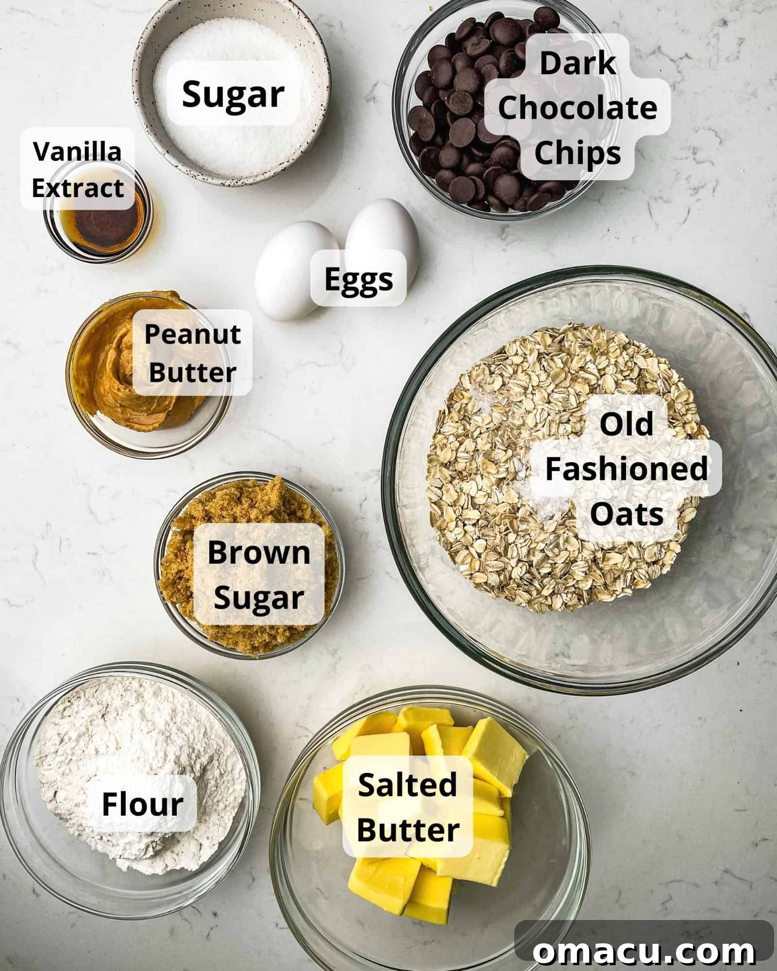 A selection of ingredients laid out on a wooden surface: old-fashioned oats, all-purpose flour, baking soda, salt, softened butter, granulated sugar, brown sugar, vanilla extract, dark chocolate chips, eggs, and creamy peanut butter.