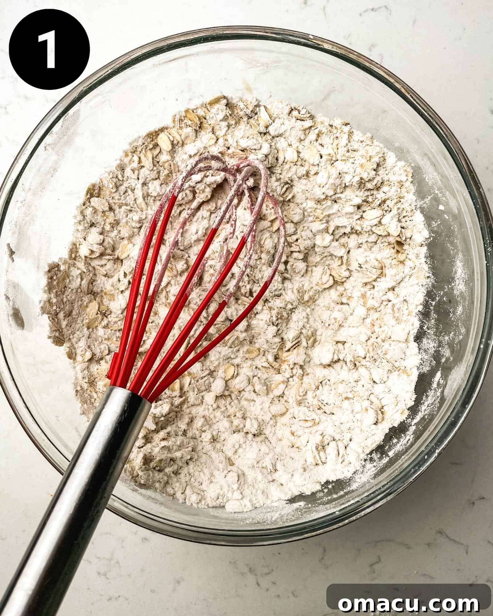 Dry ingredients, including flour, oats, baking soda, and salt, are mixed together in a large glass bowl with a whisk, ensuring even distribution.