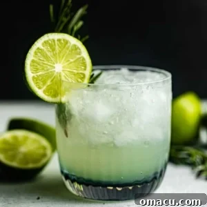 A vibrant vodka gimlet on a grey surface garnished with a lime wheel and fresh rosemary.