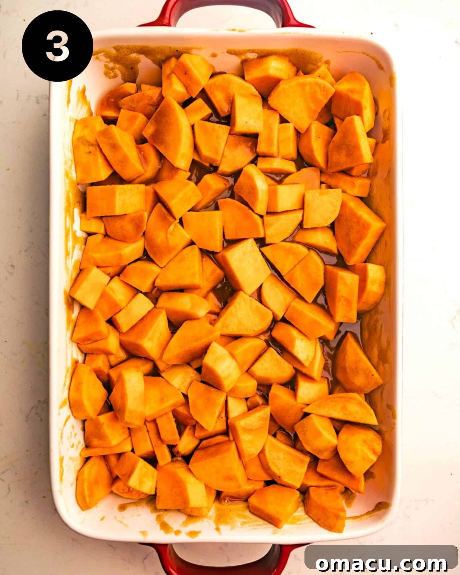A casserole dish filled with sweet potato chunks generously coated in a rich, shiny candied glaze, ready for baking.