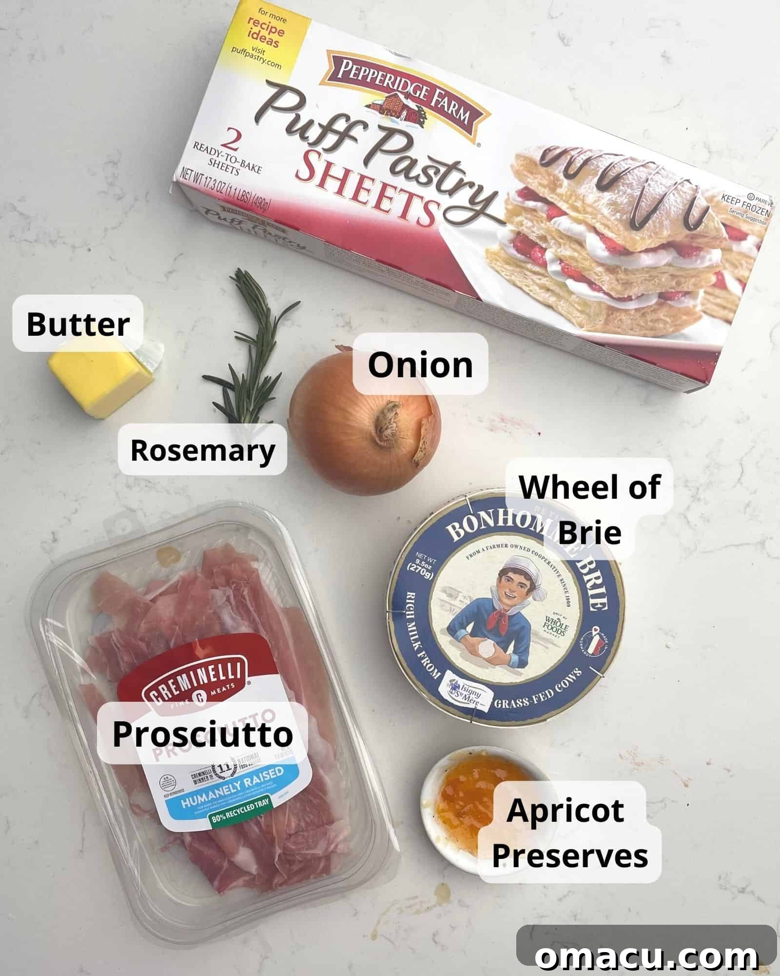 Key ingredients for baked brie en croute: puff pastry, a wheel of brie, apricot preserves, prosciutto, yellow onion, fresh rosemary, and butter.