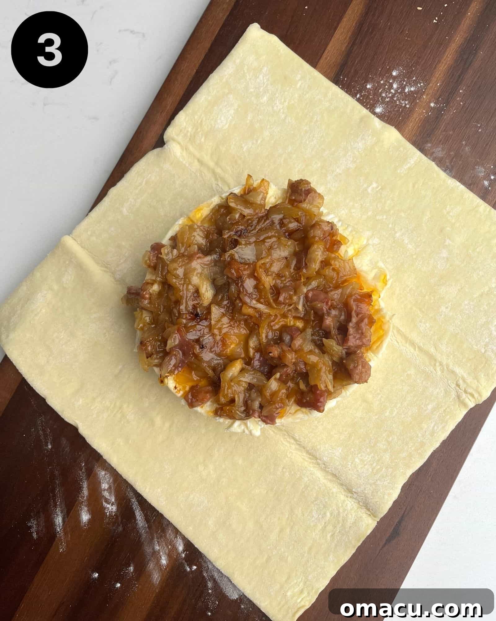 A wheel of brie beautifully topped with apricot preserves, caramelized onions, prosciutto, and fresh rosemary, ready to be enclosed in puff pastry.