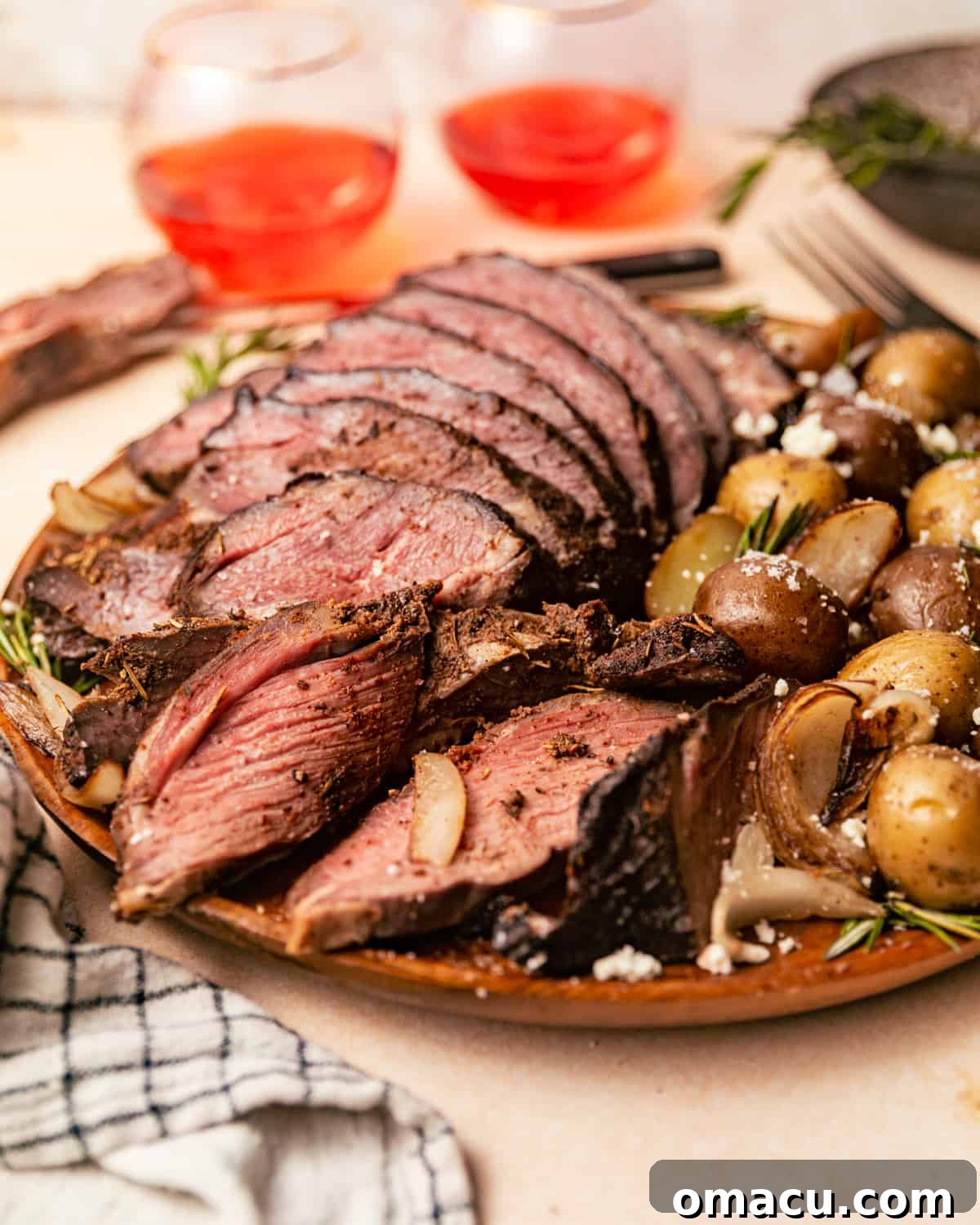Sliced smoked leg of lamb on a wooden plate with roasted potatoes and fresh herbs, ready to be served.
