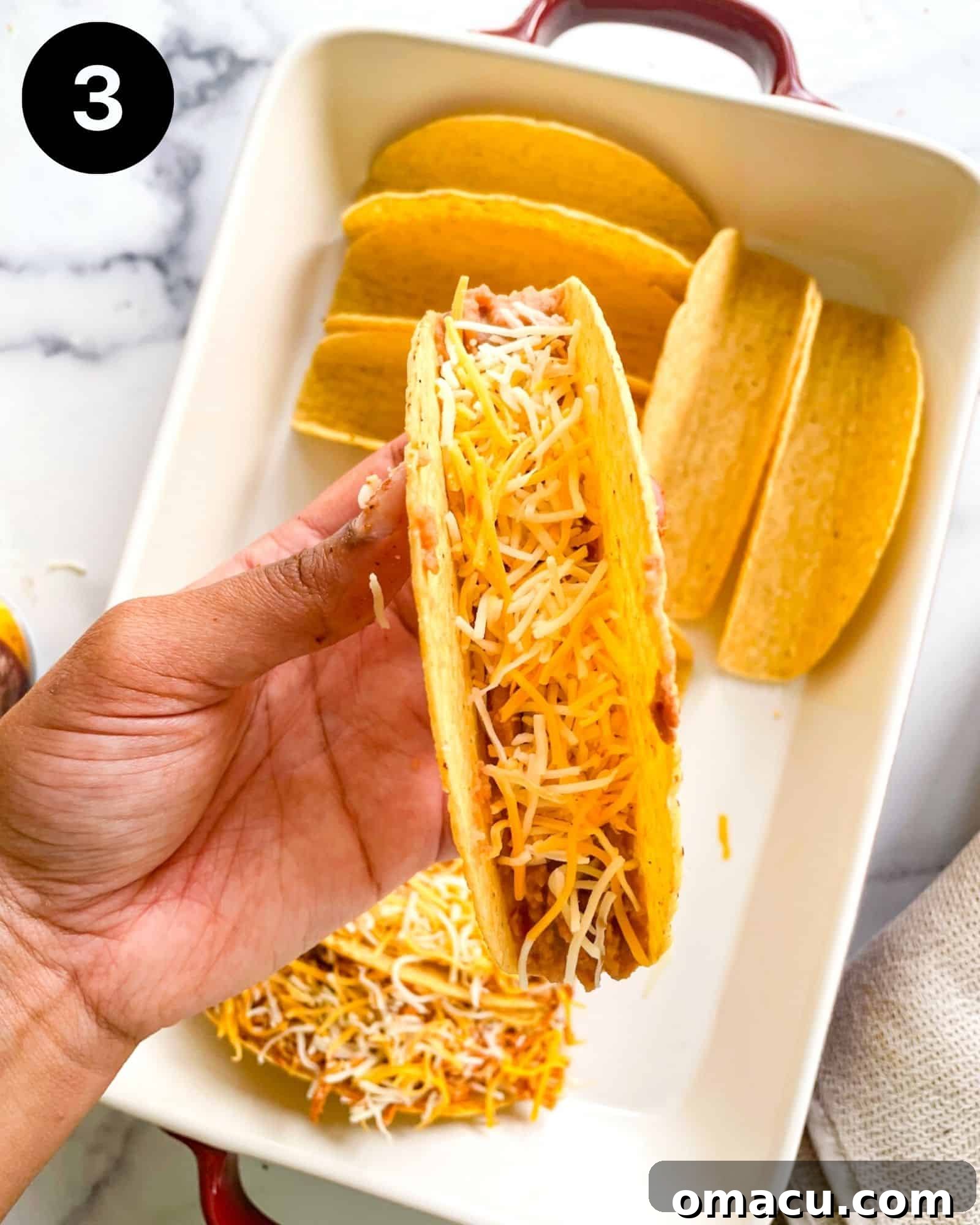A close-up shot of a hard taco shell being carefully filled with creamy refried beans and a layer of shredded cheese, demonstrating the first steps of taco assembly.