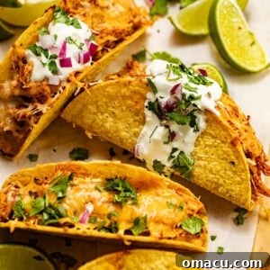 baked chicken tacos on a piece of parchment paper topped with cheese, sour cream, and diced red onions.
