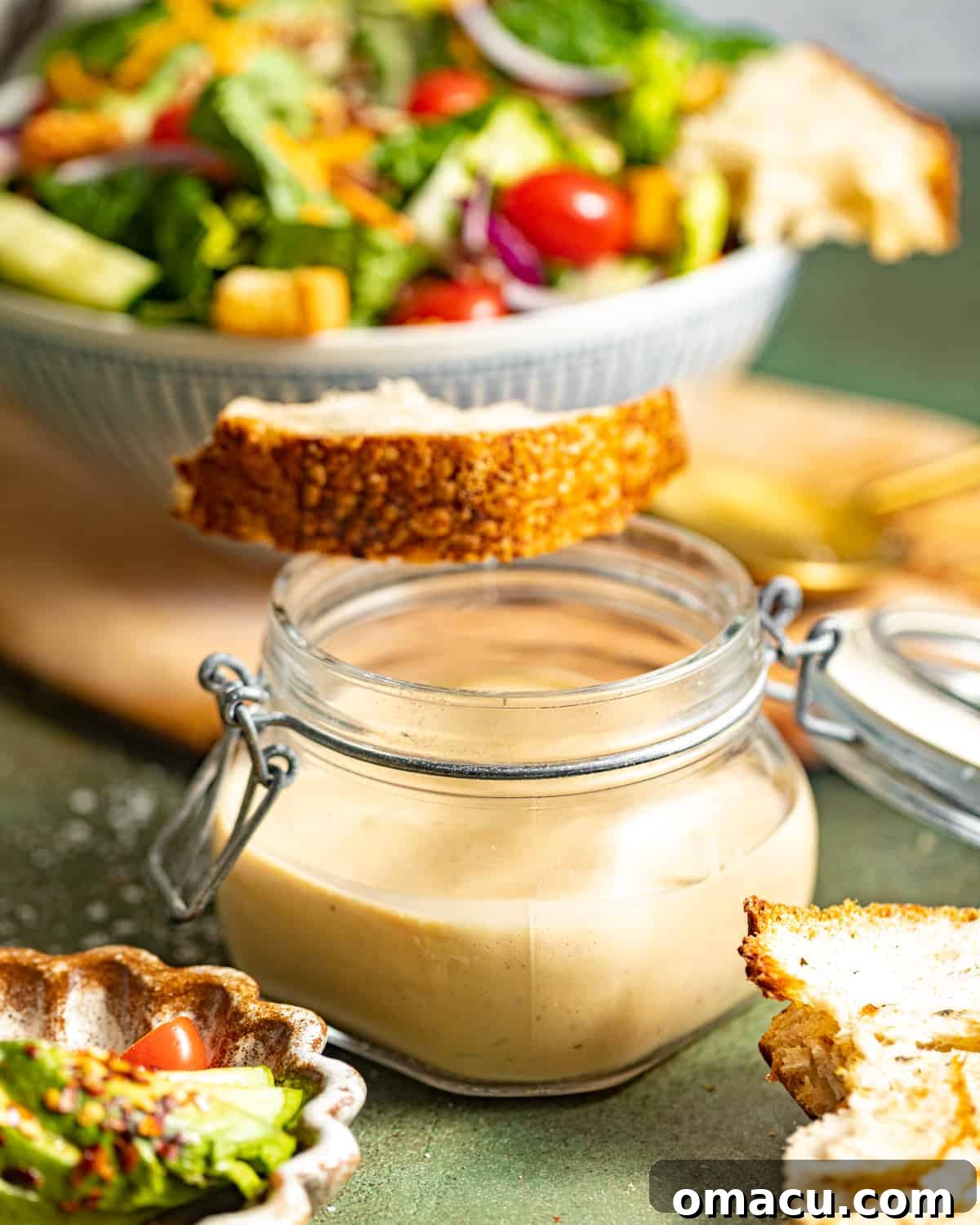 A jar of rich, homemade creamy balsamic dressing in front of a fresh, green salad topped with a slice of artisan bread.