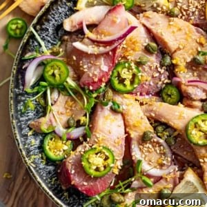 A plate of exquisite Hamachi Crudo, thinly sliced yellowtail arranged with serrano peppers, sesame seeds, red onions, and capers for a burst of fresh flavor.