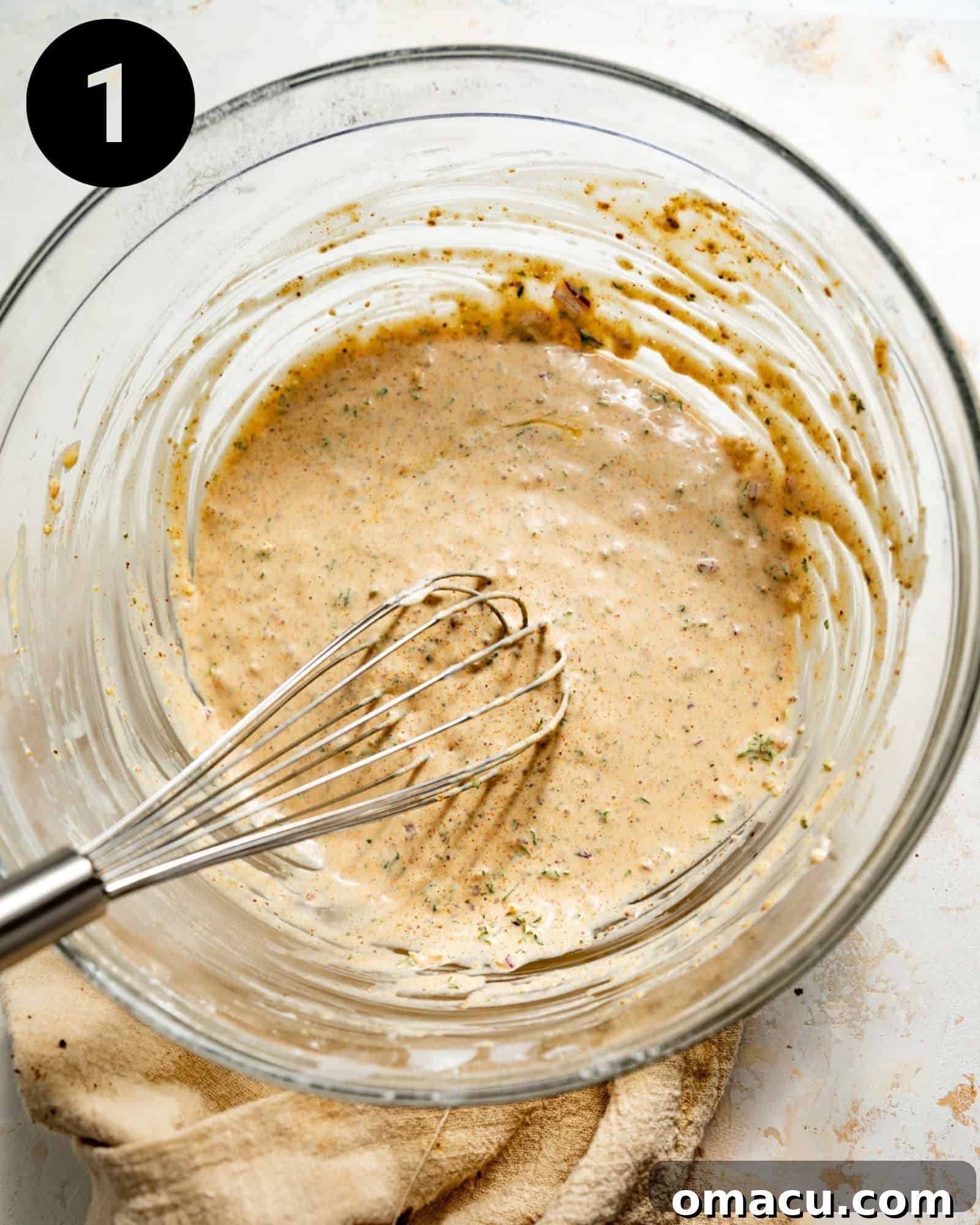 A whisk stirring a creamy mixture of mayo and seasonings in a glass mixing bowl, forming the base for delicious crab cakes.