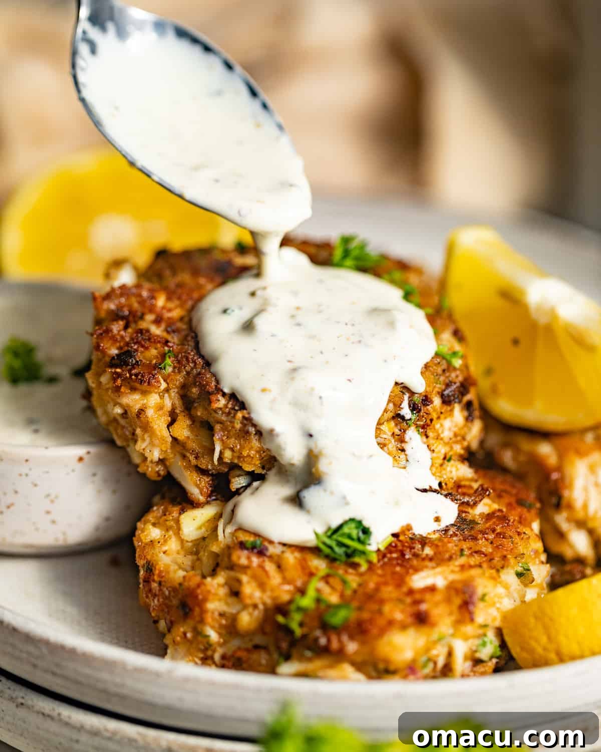 Homemade tartar sauce drizzling off a spoon onto a stack of crispy pan-fried crab cakes, with fresh lemon.