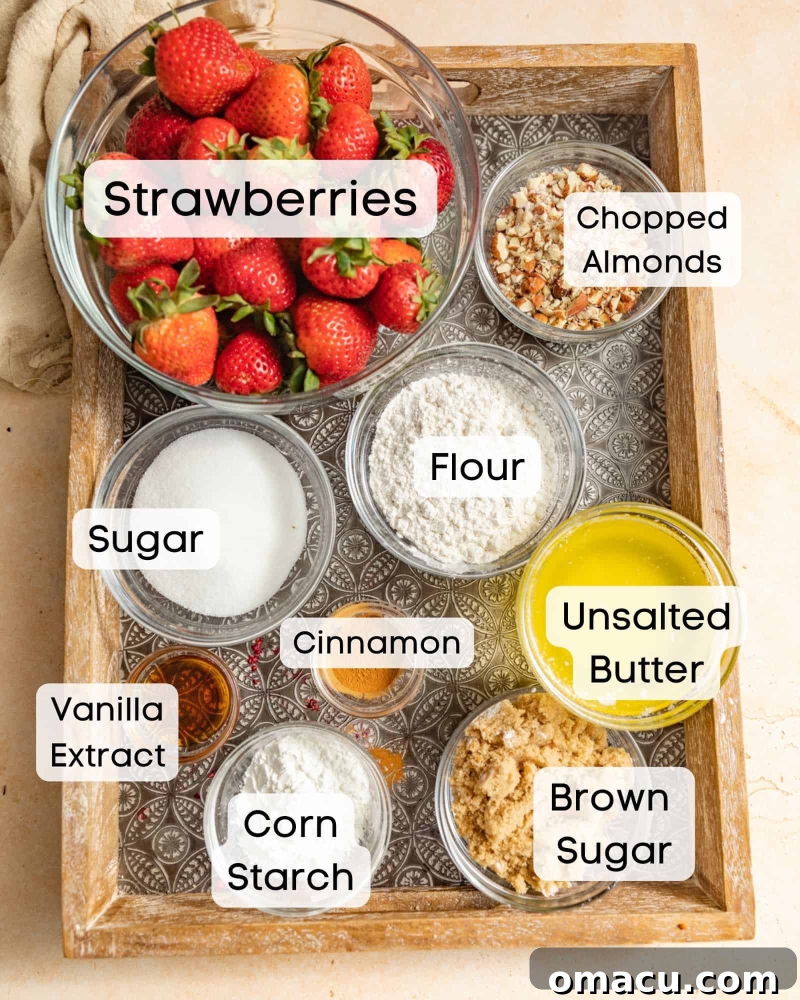 Various ingredients laid out for making strawberry crumble: fresh strawberries, brown sugar, white sugar, unsalted butter, corn starch, lemon juice, all-purpose flour, vanilla extract, cinnamon, and chopped almonds.