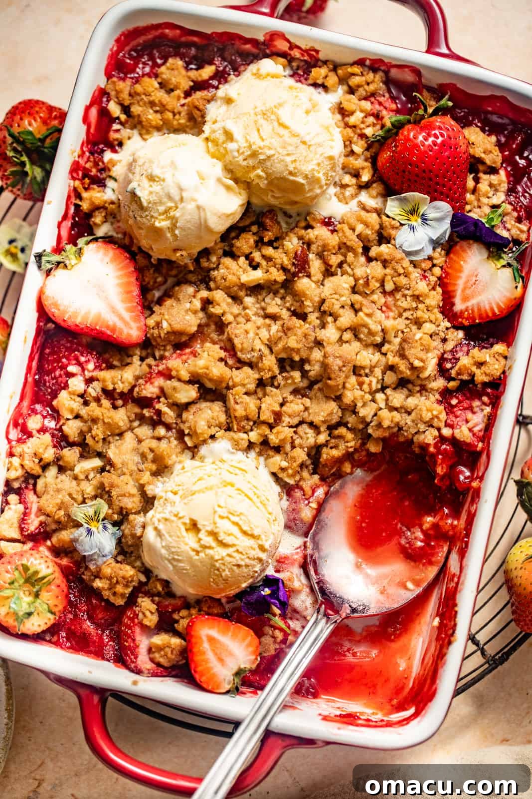 A baked strawberry crisp in a square baking dish, topped with ice cream and a spoon, ready to be served.