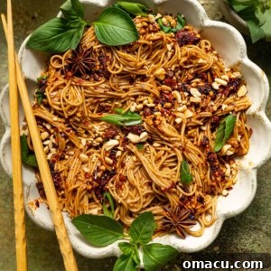 Chopsticks resting on a white scalloped bowl filled with spicy peanut noodles.