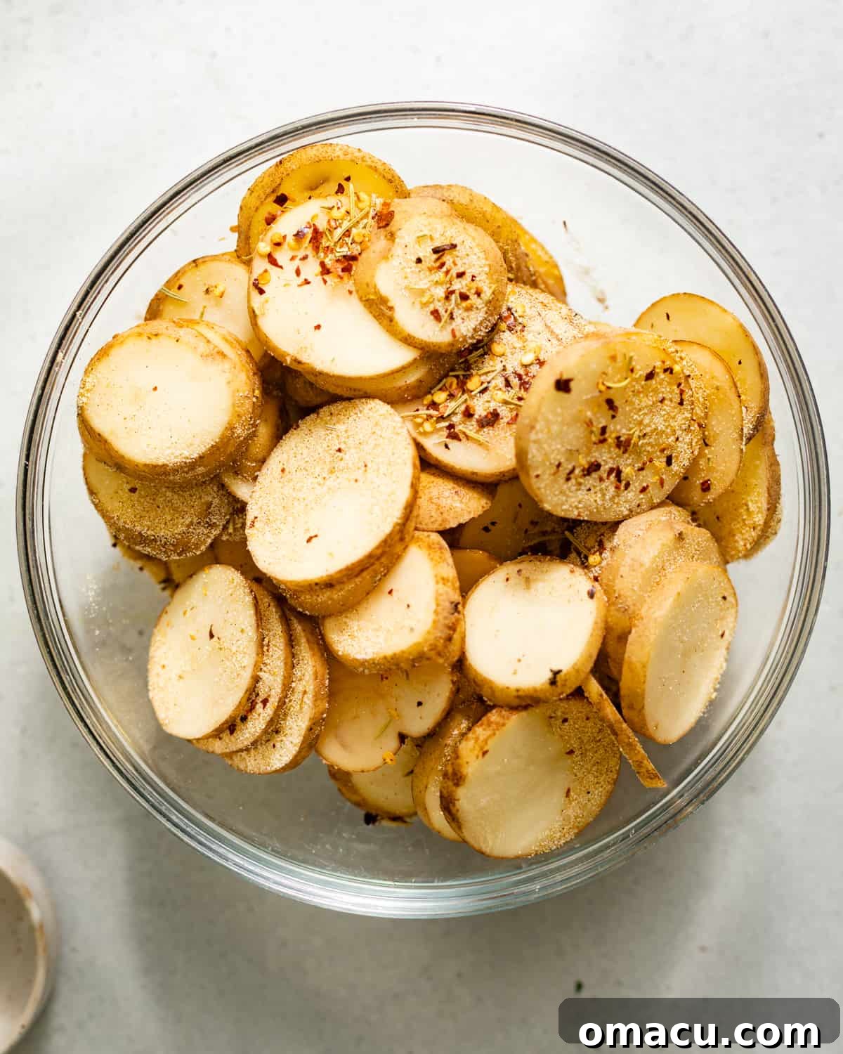 Sliced potato rounds with seasoning sprinkled on in a glass mixing bowl, ready for frying.