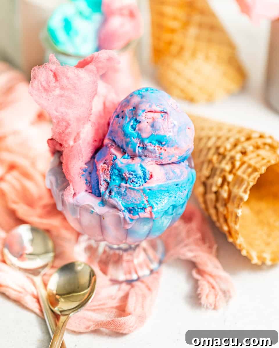 A vibrant swirl of pink and blue cotton candy ice cream in an elegant glass serving cup, flanked by two spoons and crisp waffle cones in the background, inviting a taste of carnival nostalgia.