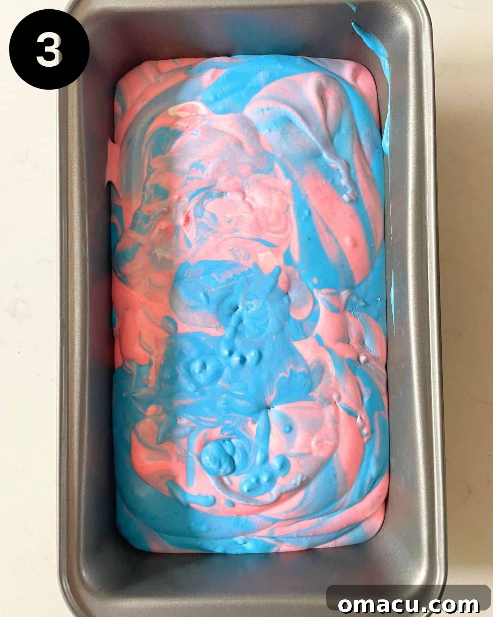 A loaf pan filled with beautifully swirled pink and blue cotton candy ice cream mixture, ready for the freezer.