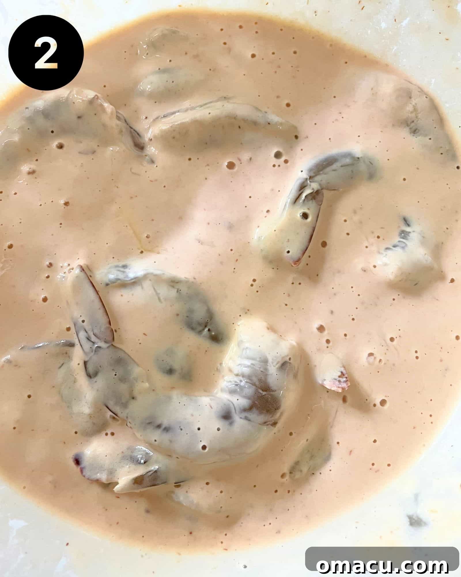 Raw shrimp marinating in a creamy, orange-hued Boom Boom sauce mixture in a glass bowl.