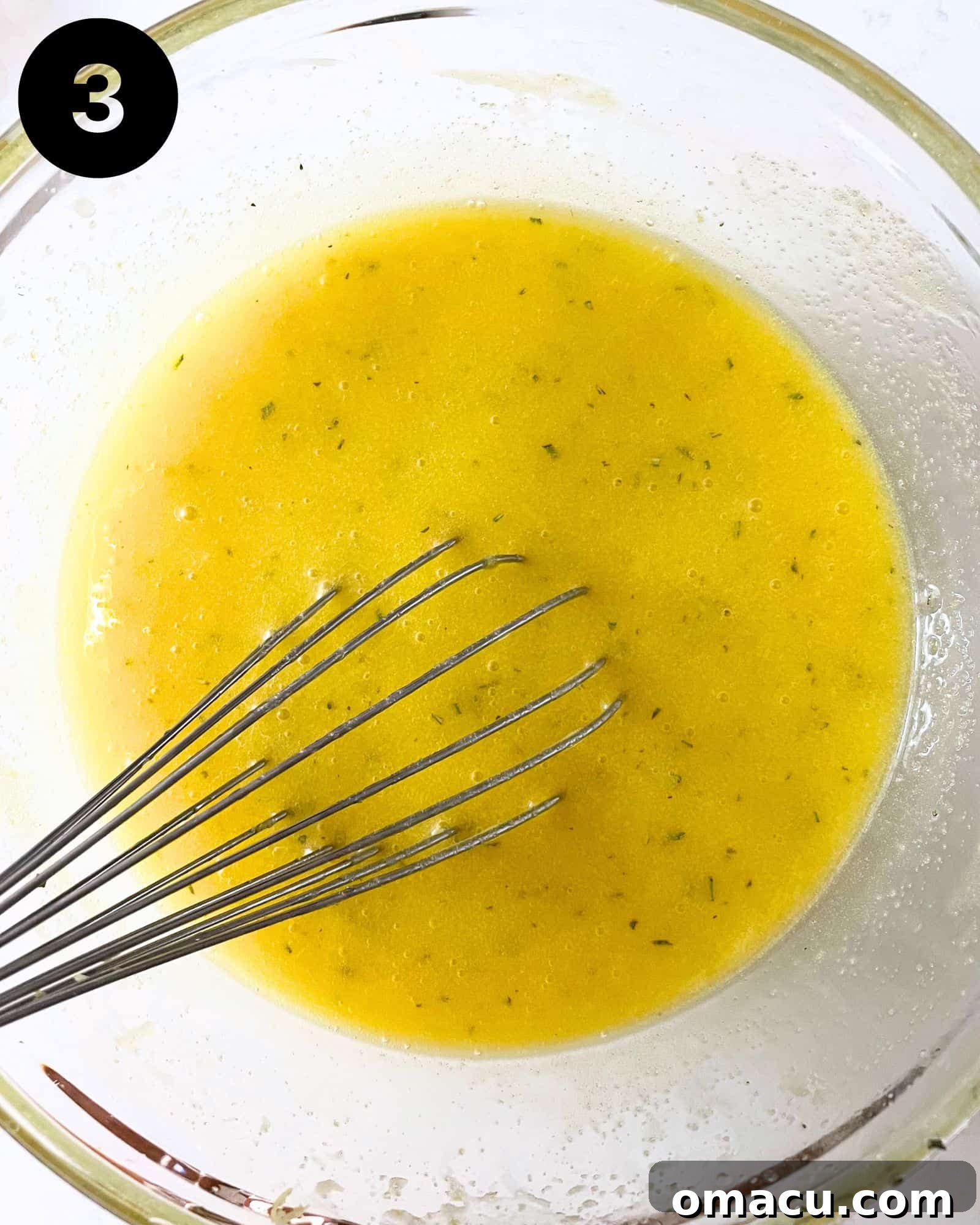 Olive oil, sugar infused with lemon and rosemary, eggs, and lemon juice are whisked together in a bowl, creating a smooth wet mixture.