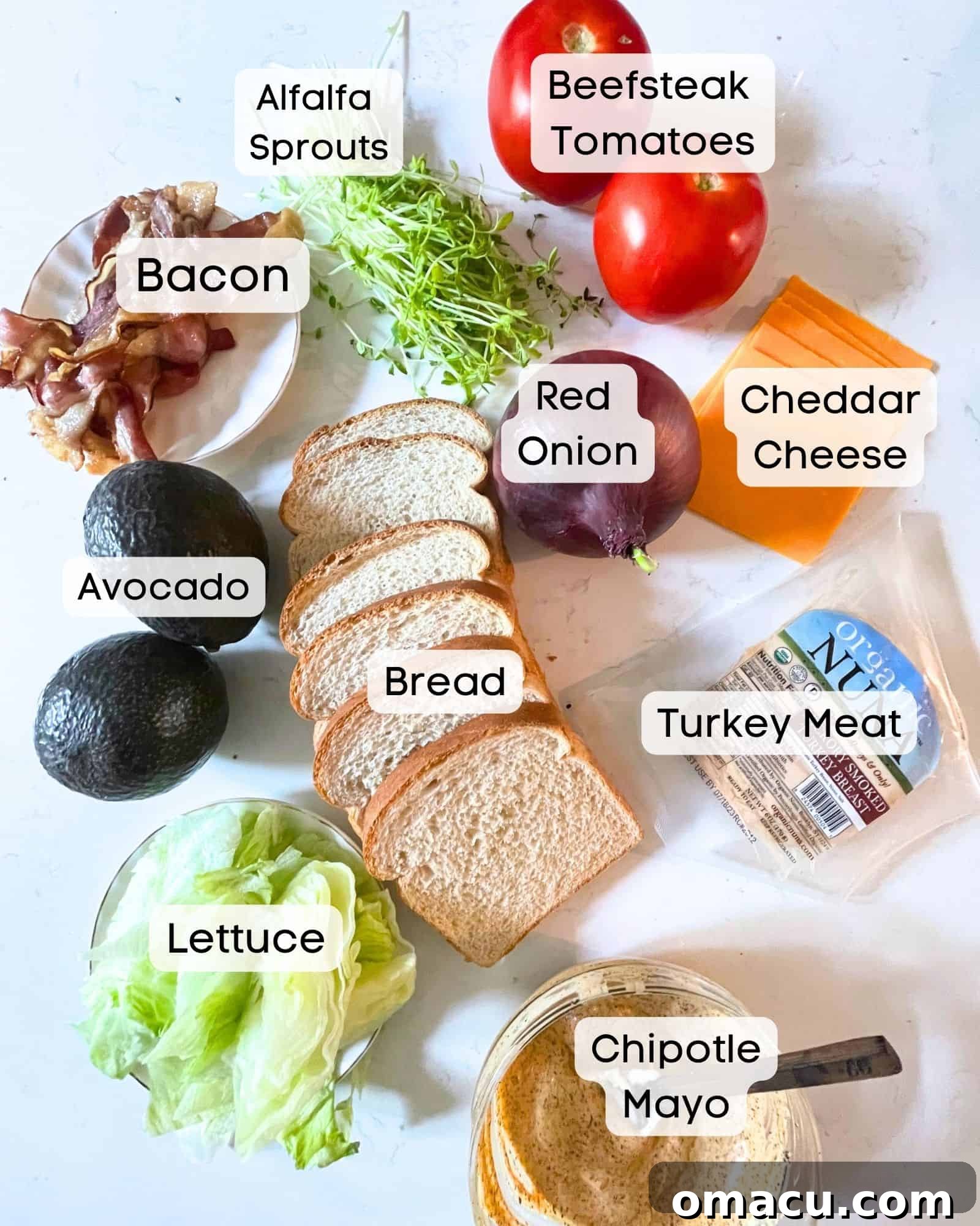 Ingredients laid out on a white surface for making a California Club Sandwich: bread, chipotle mayo, tomato, lettuce, avocado, cheddar cheese, red onion, sliced turkey, crispy bacon, and alfalfa sprouts.