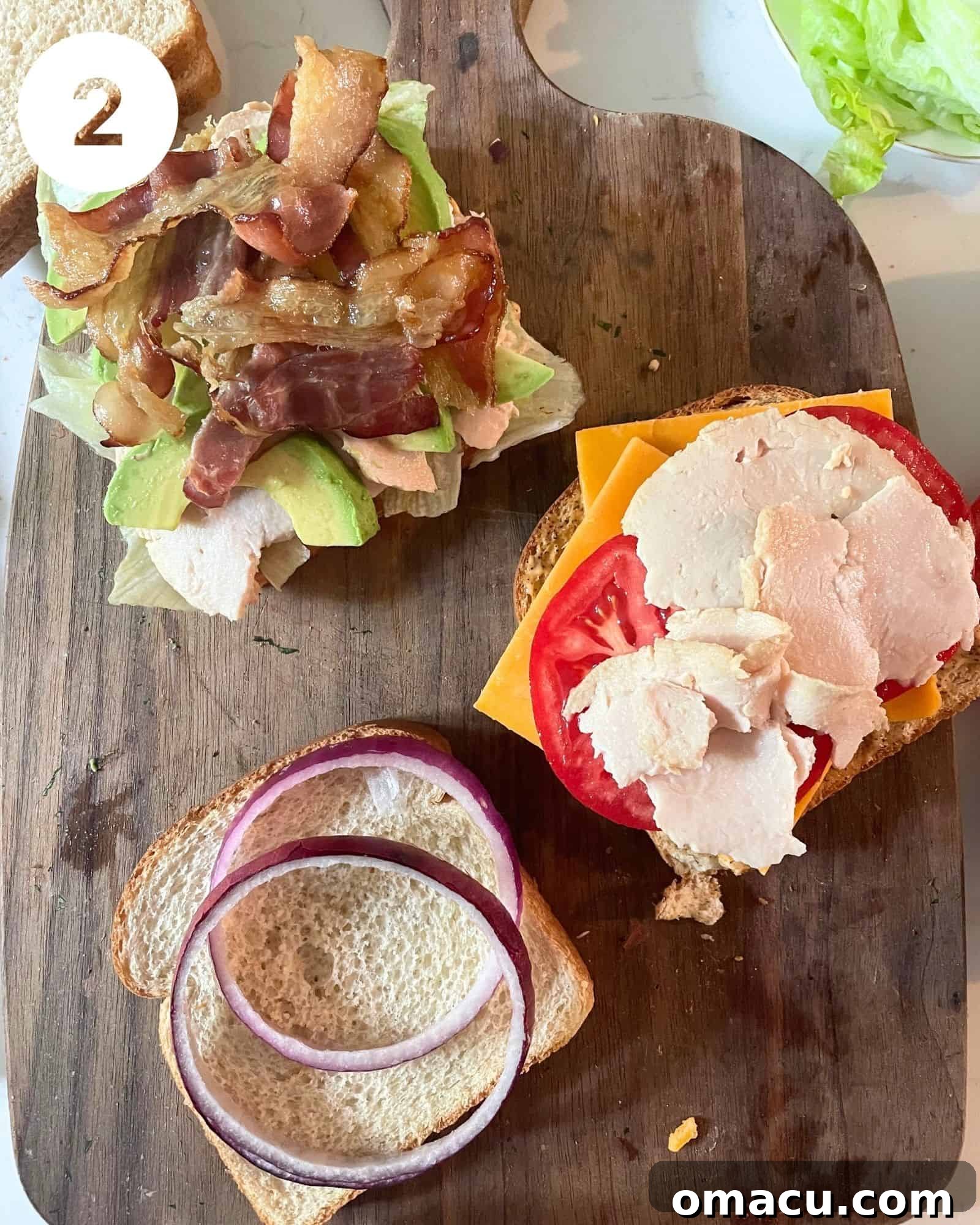 The first layer of a California Club Sandwich being assembled with toasted bread, cheese, turkey, bacon, and red onion.