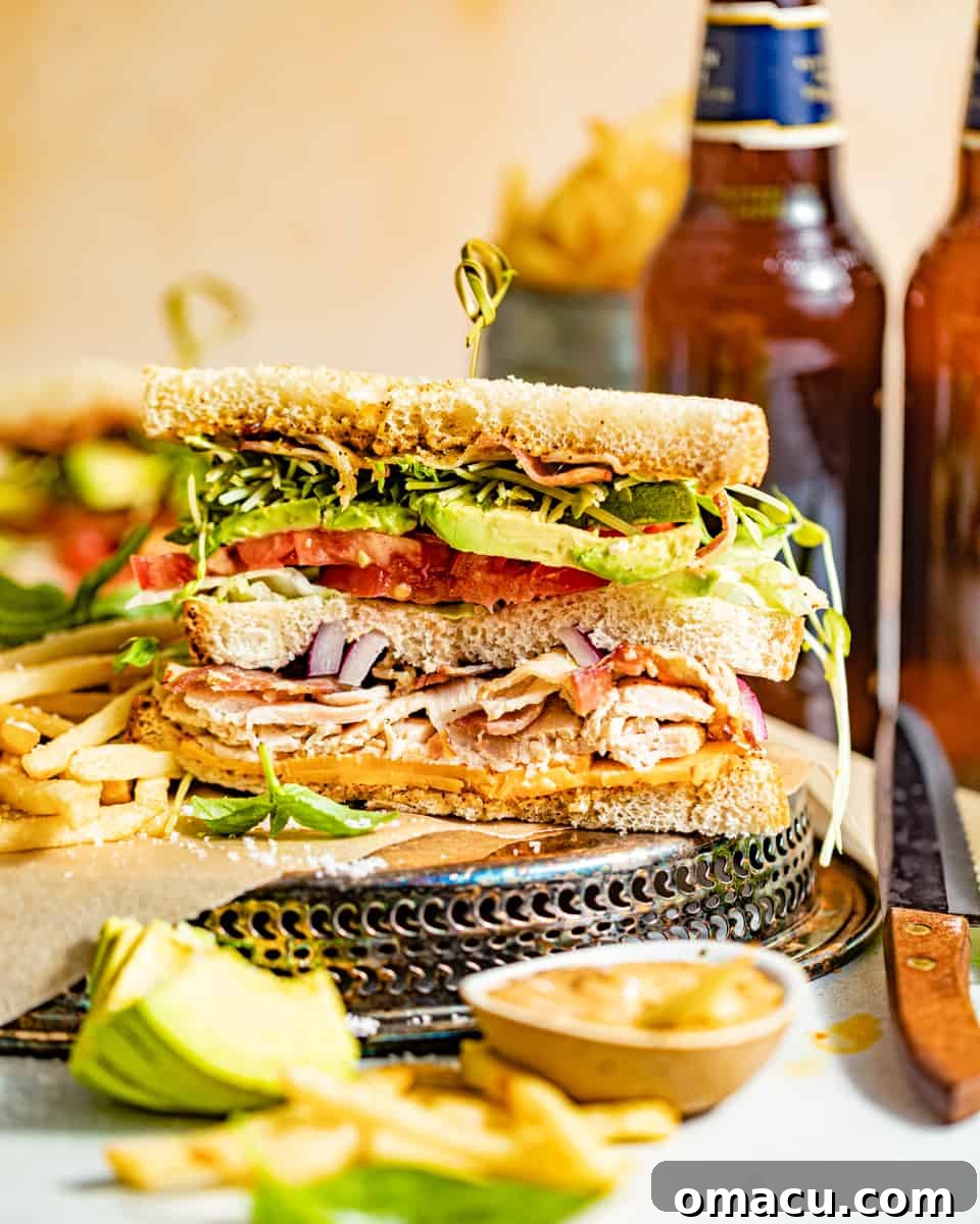 A California Club Sandwich, cut in half, presented on a plate with two cans of soda in the background and sliced avocado in the foreground.