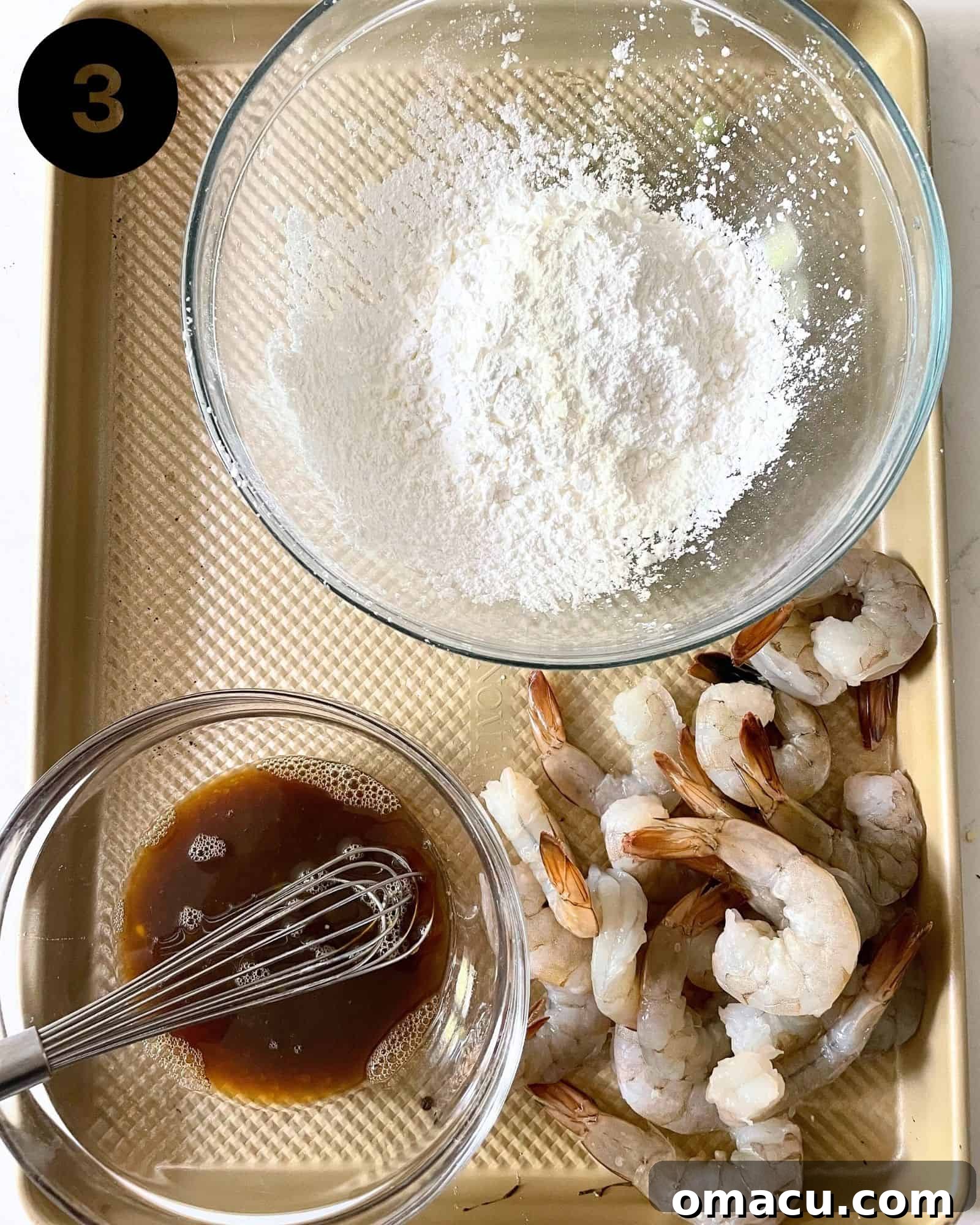The breading station setup: a bowl of cornstarch, a bowl with Shaoxing wine, soy sauce, and egg mixture, and raw shrimp on a baking sheet, ready for preparation.