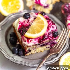 A perfectly frosted and drizzled slice of lemon blueberry cake on a plate, garnished with fresh lemon and blueberries.