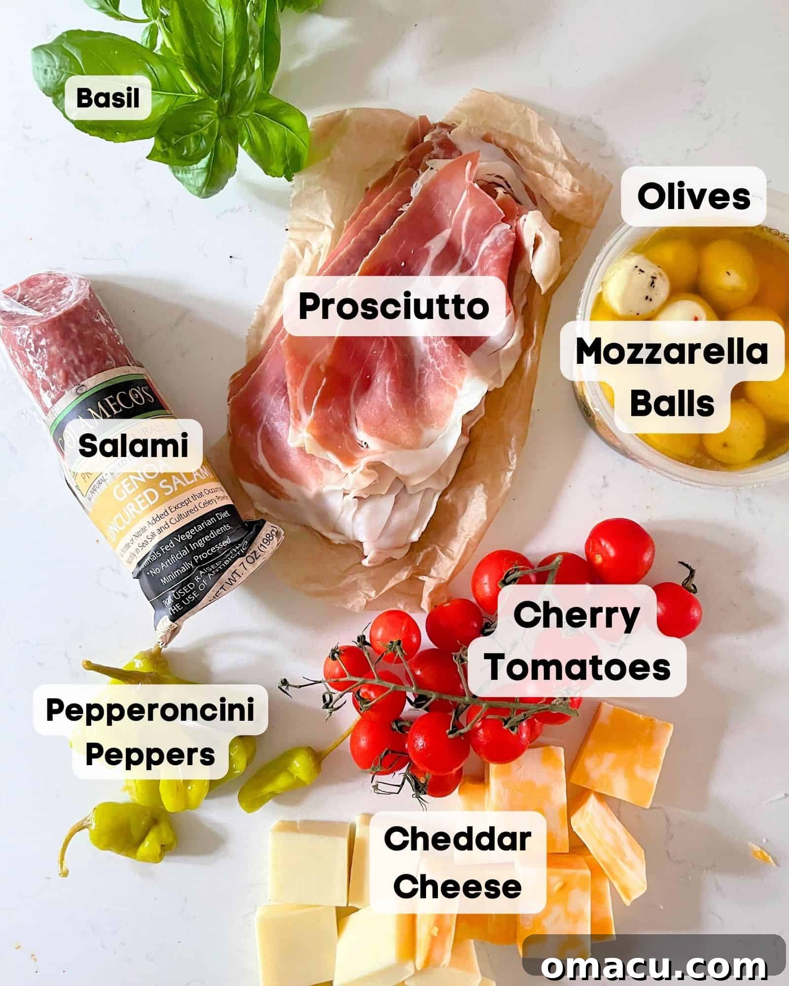 A selection of fresh ingredients including prosciutto, cheddar cheese, basil, pepperoncini peppers, salami, cherry tomatoes, olives, and mozzarella balls, laid out for making charcuterie skewers.