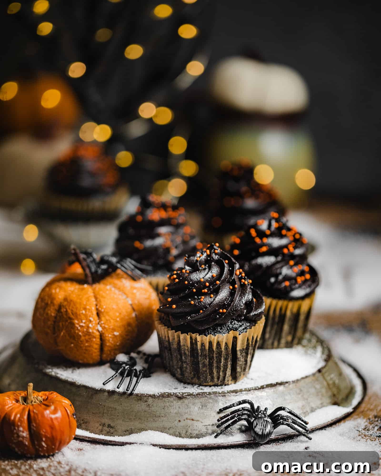 Three black velvet cupcakes on a plate with a pumpkin and fake spiders, set against a dark, festive background.