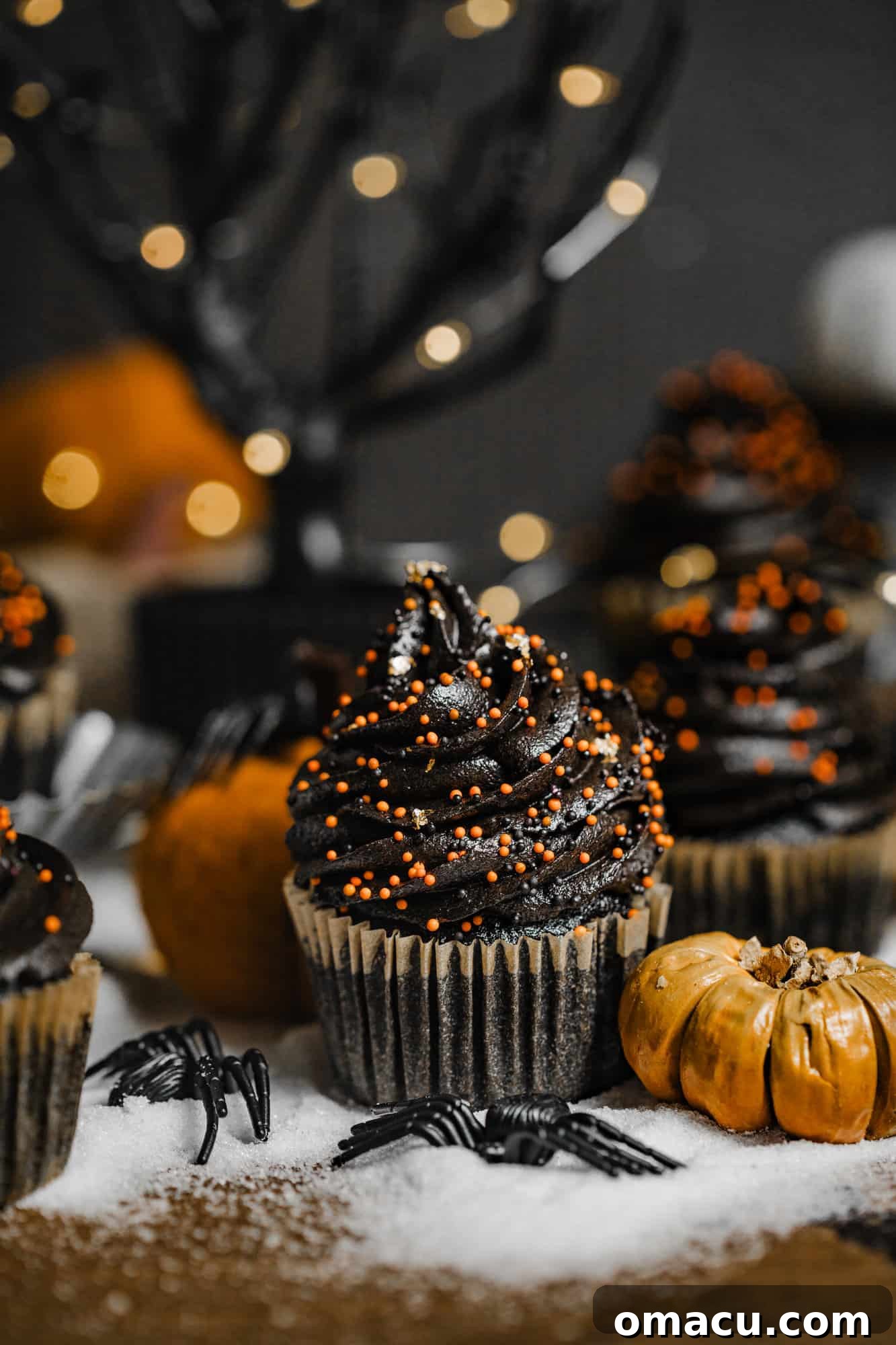 A black velvet cupcake in front of a black tree with pumpkins and fake spiders, perfectly decorated.