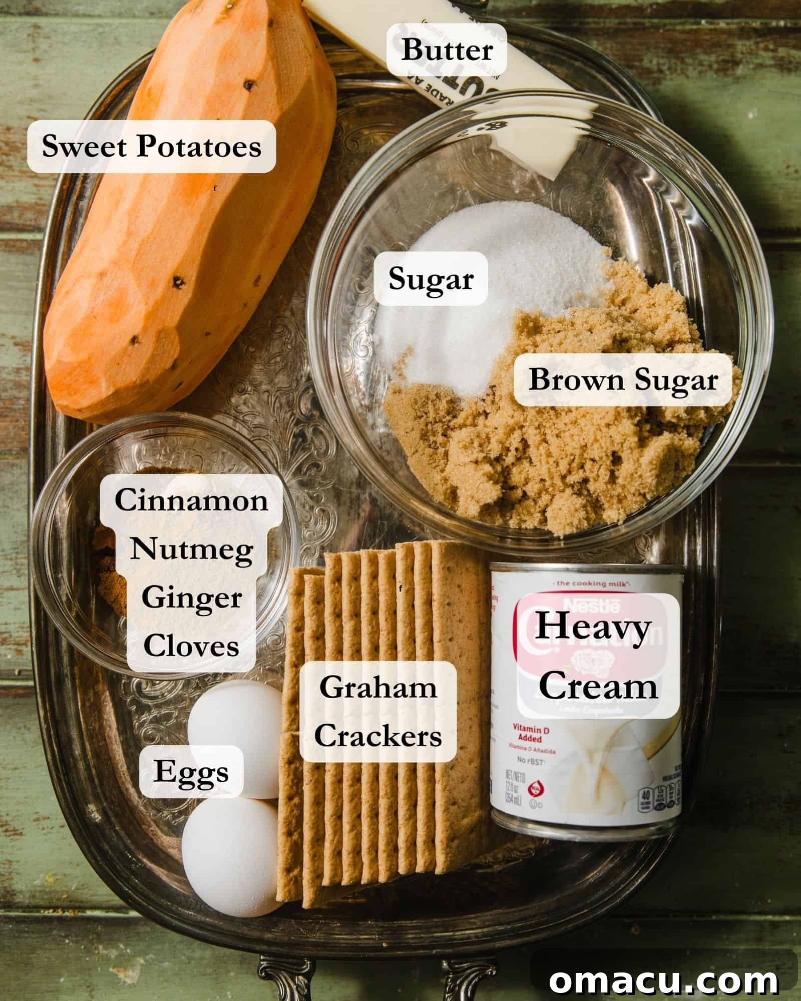 ingredients to make sweet potato pie on a silver tray - butter, sweet potatoes, sugar, brown sugar, heavy cream, graham crackers, eggs, cinnamon, ginger, and nutmeg.