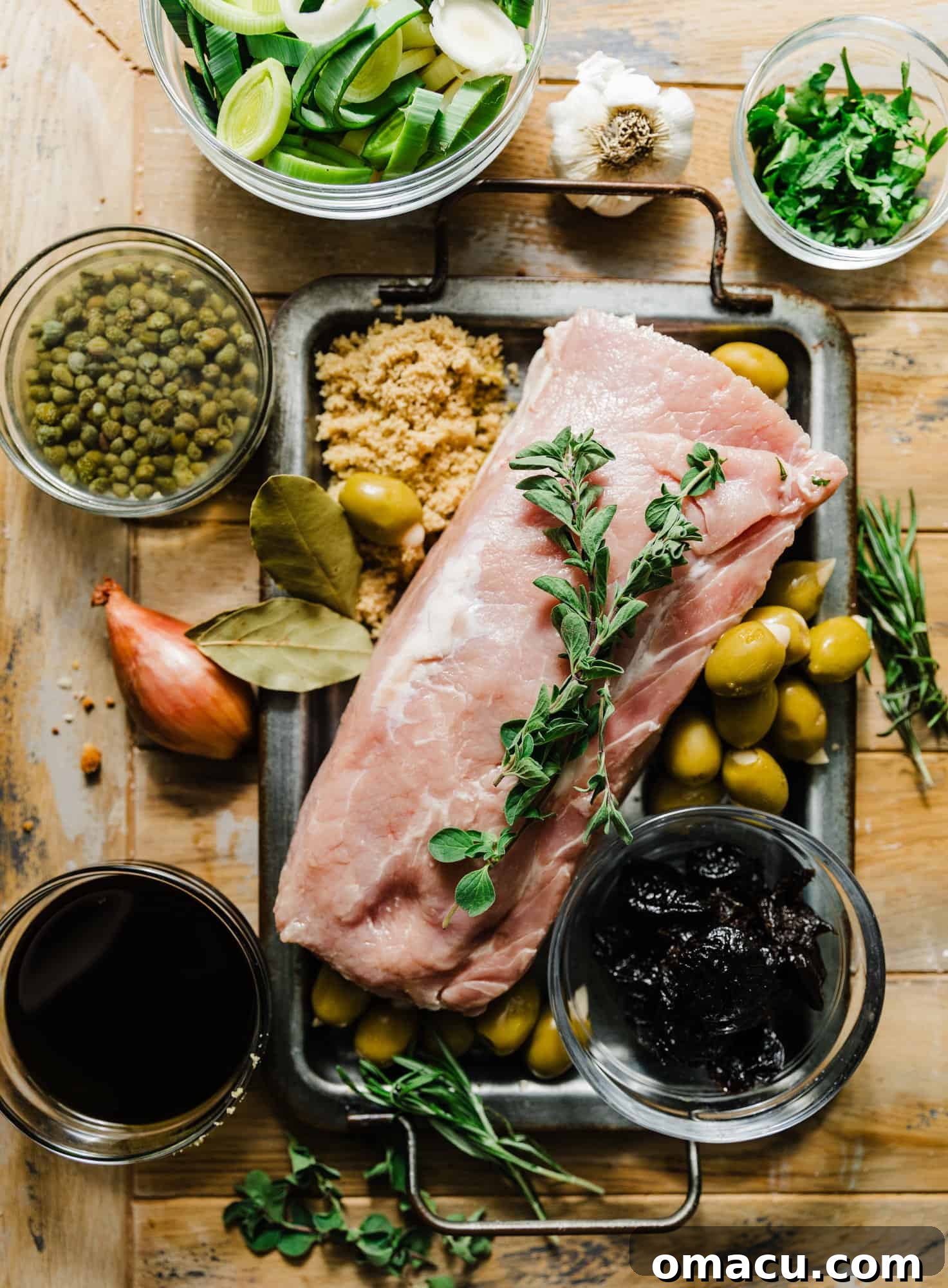 A vibrant flat lay of ingredients for Pork Marbella, including pork tenderloin, fresh herbs like oregano and rosemary, shallots, prunes, green olives, olive oil, balsamic vinegar, brown sugar, garlic, and leeks.