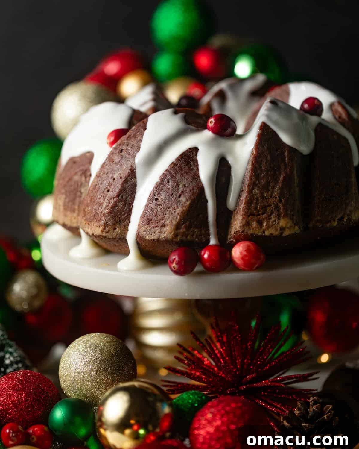 A beautifully decorated gingerbread bundt cake on a white cake stand, artfully drizzled with cream cheese frosting and surrounded by festive Christmas decorations.