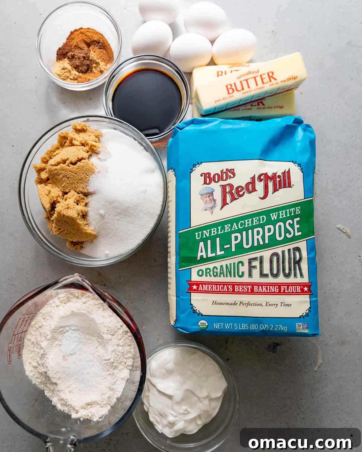 A clean, organized display of all the dry and wet ingredients needed to bake a gingerbread bundt cake, arranged on a white surface.