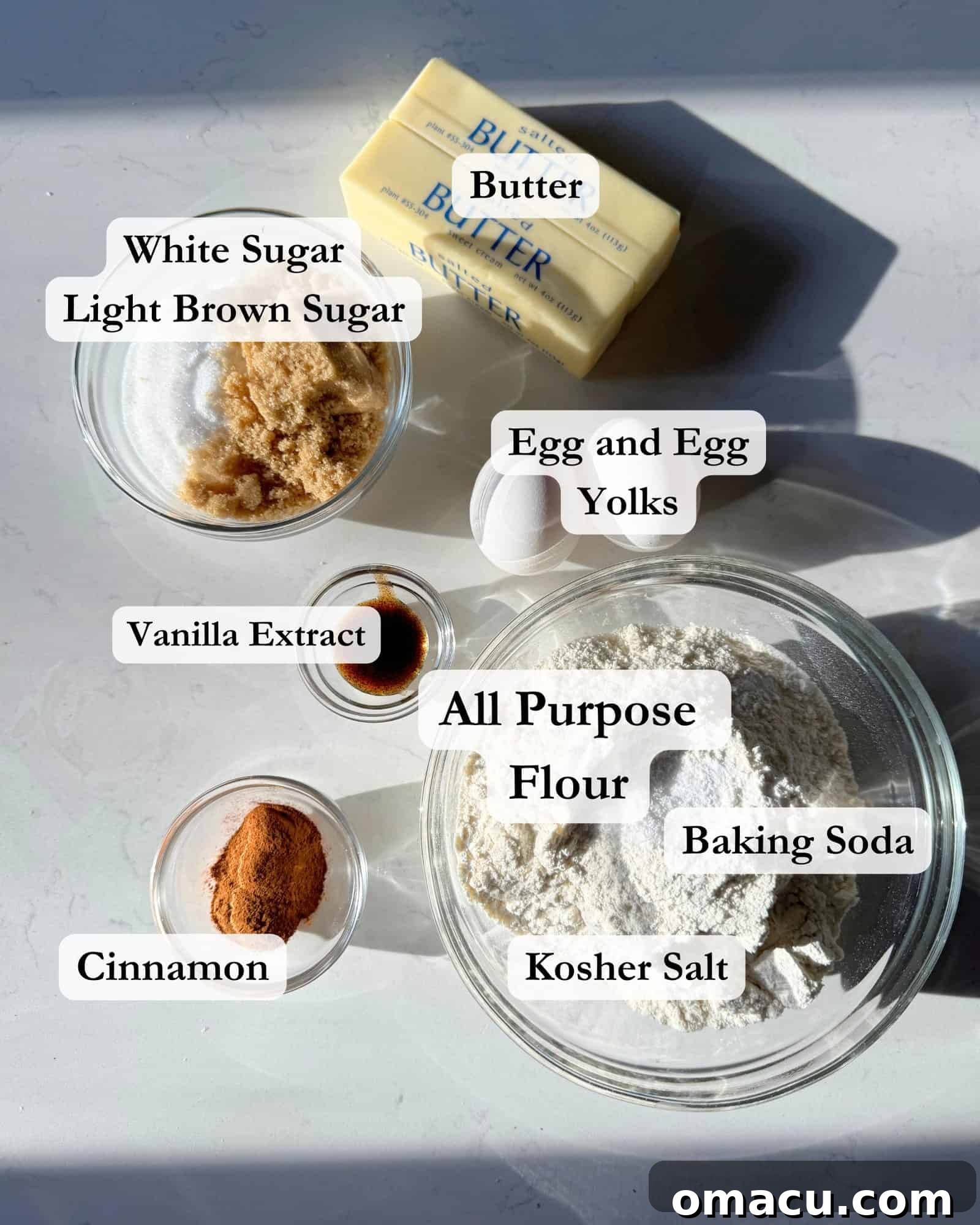 Bowls of ingredients including flour, cinnamon, baking soda, salt, brown sugar, white sugar, eggs, butter, egg yolk, and vanilla extract, arranged on a white surface for making cinnamon cookies