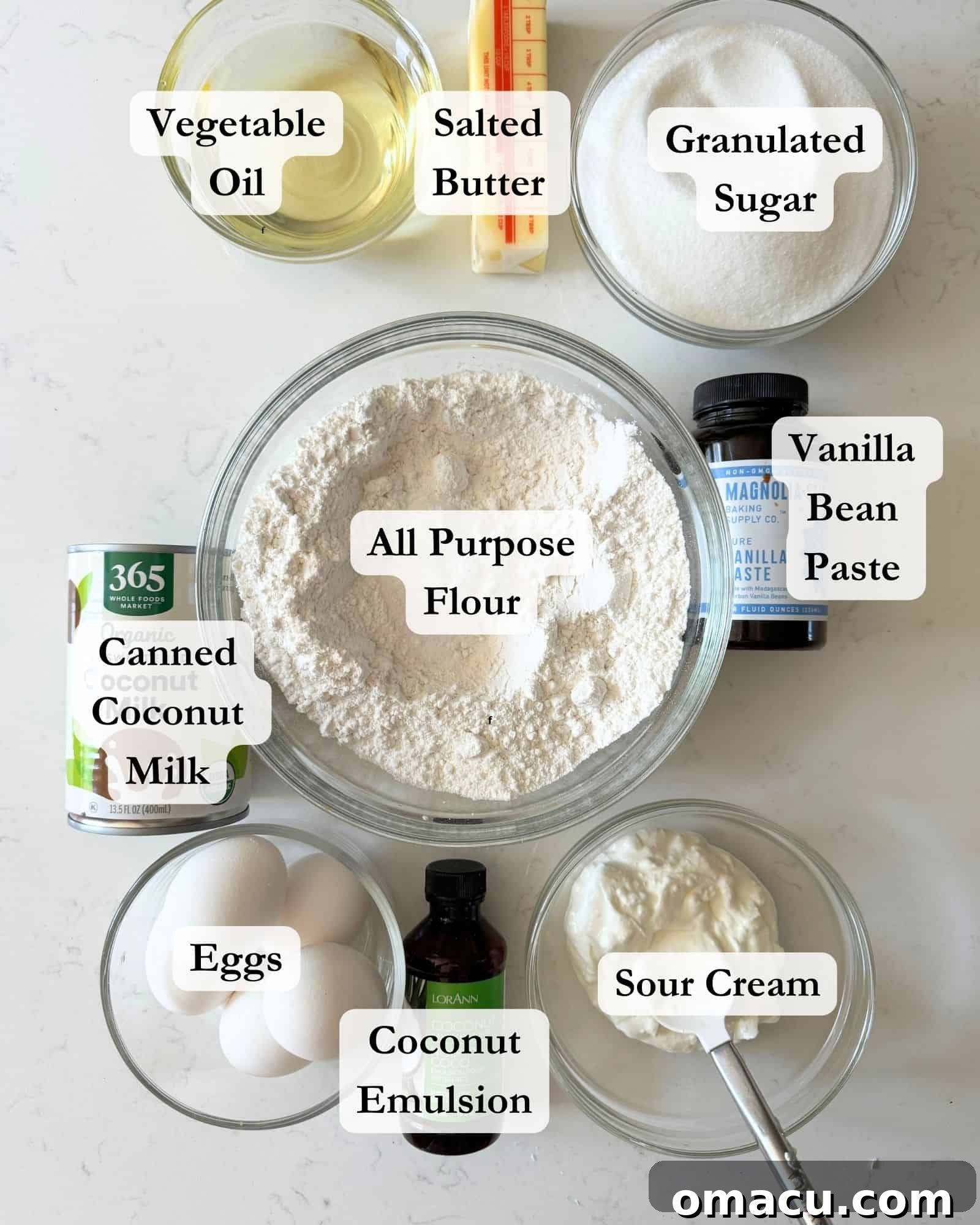 A collection of key ingredients for making a coconut layer cake, arranged in glass bowls on a white surface. Ingredients include cake flour, sugar, eggs, baking powder, kosher salt, sour cream, coconut emulsion, butter, vegetable oil, and vanilla extract.