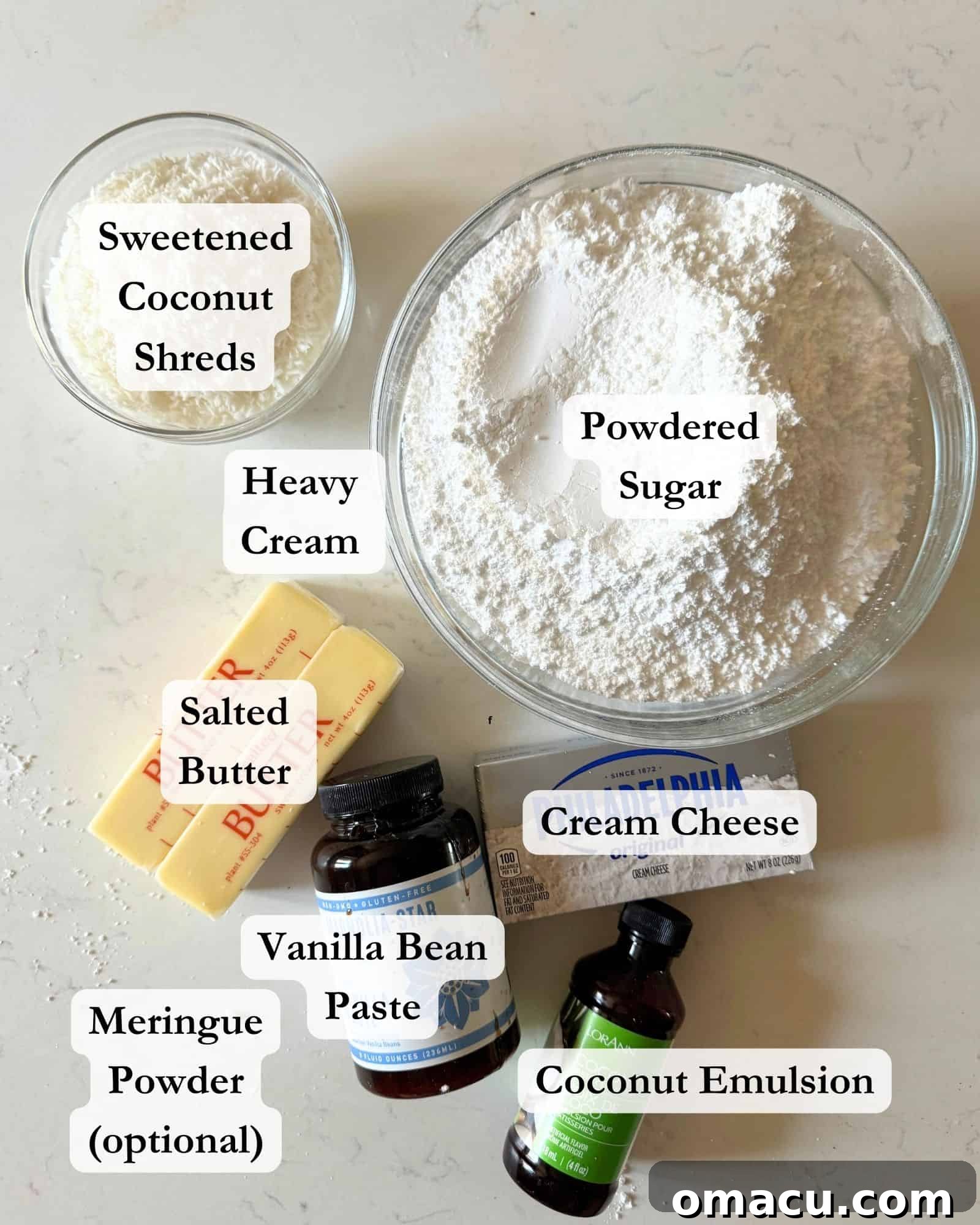 Ingredients for coconut cream cheese frosting displayed on a white surface, including powdered sugar, cream cheese, butter, meringue powder, coconut emulsion, vanilla bean paste, and heavy cream.