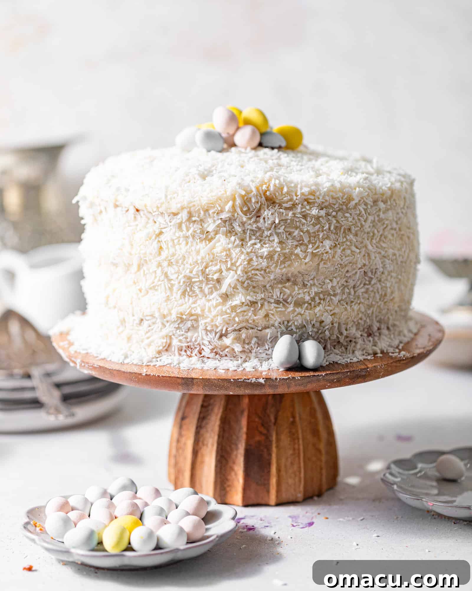 A fully assembled coconut cake on a cake stand, flanked by serving plates and bowls, ready for a gathering.
