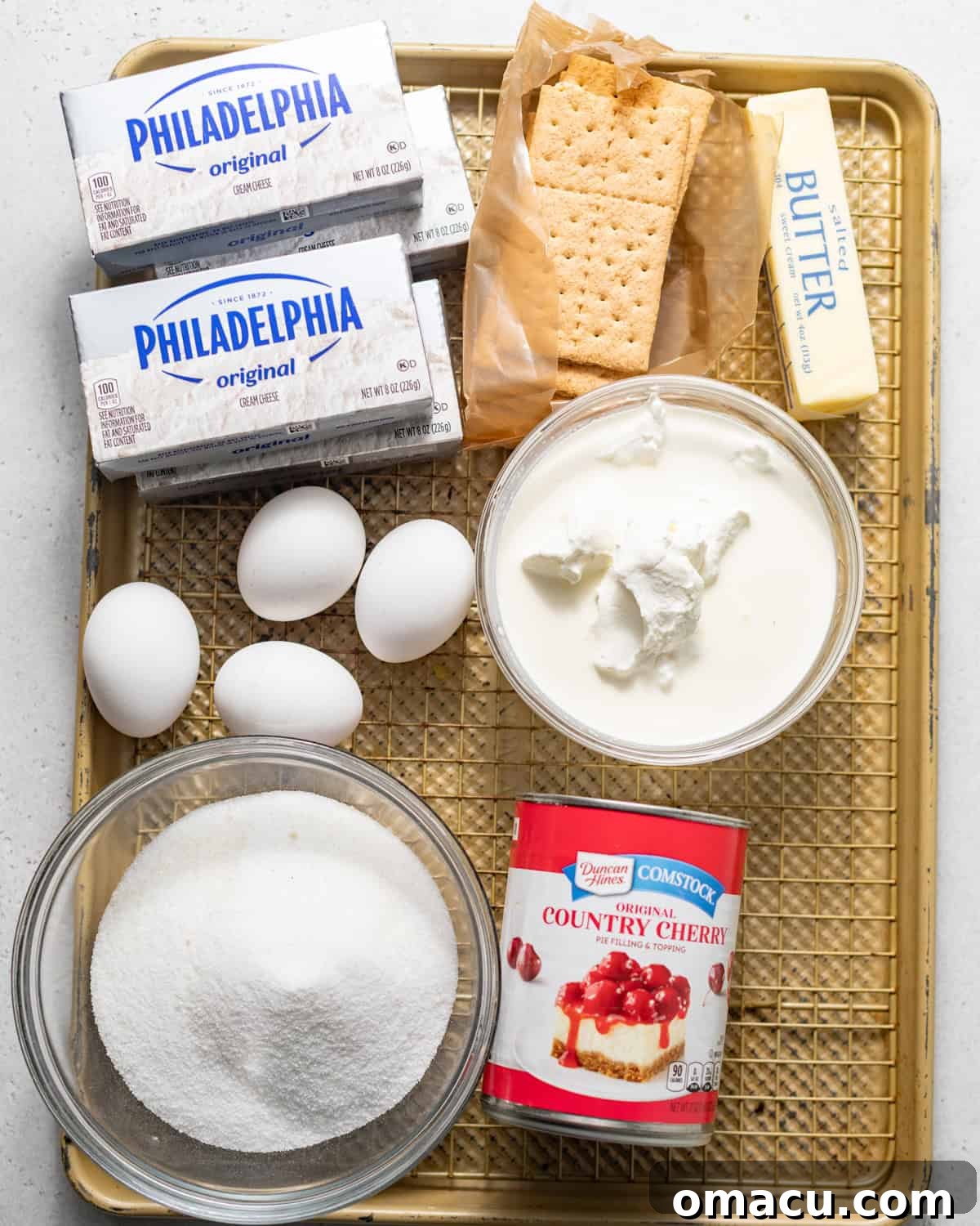 A selection of ingredients laid out on a baking tray, ready to make cherry cheesecake: cream cheese, sugar, eggs, cornstarch, vanilla extract, heavy cream, sour cream, graham crackers, butter, and a can of cherry pie filling.