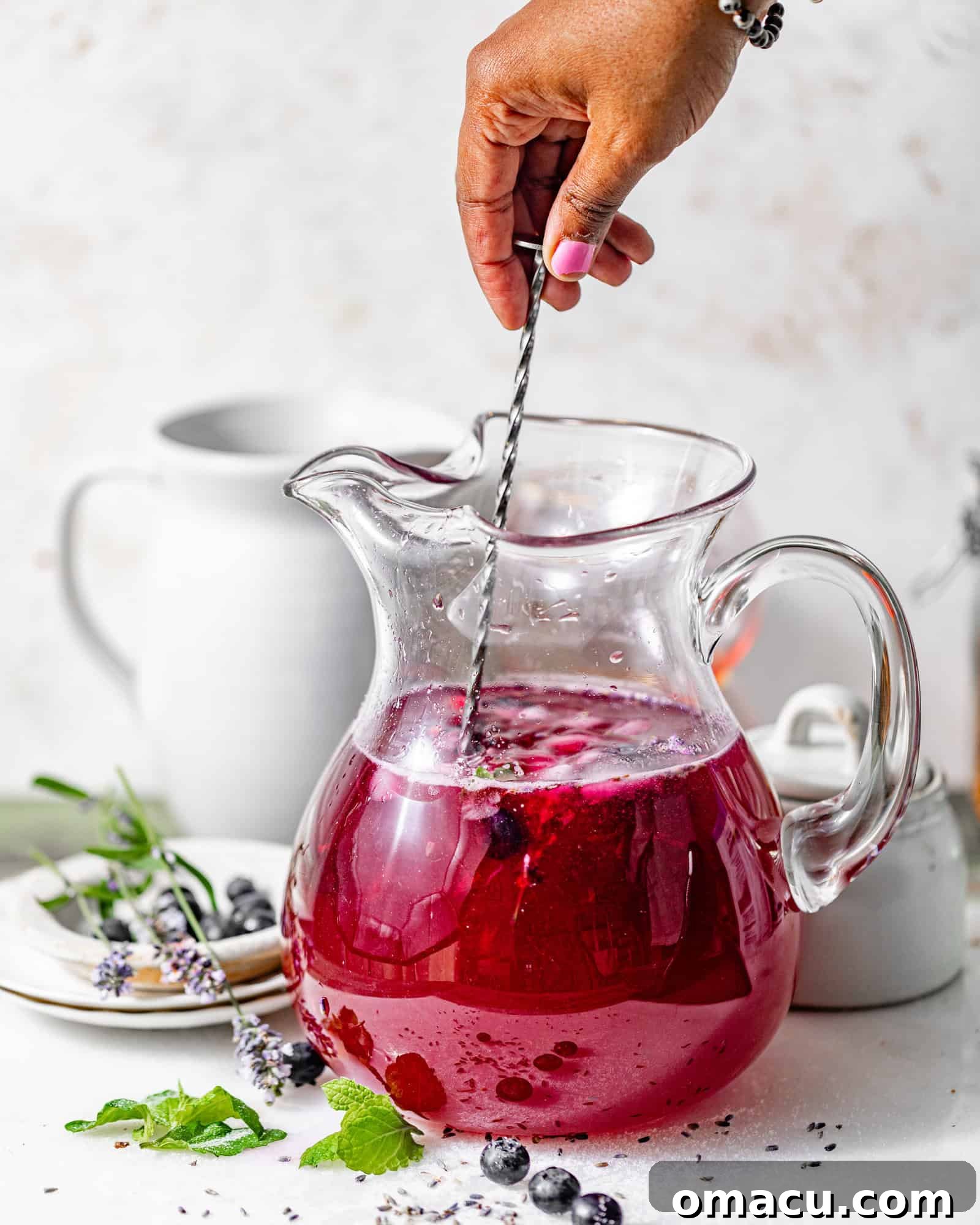A hand stirring the vibrant blueberry lavender syrup into a pitcher of fresh lemonade with a long spoon, creating a beautiful swirling effect of purple and yellow.