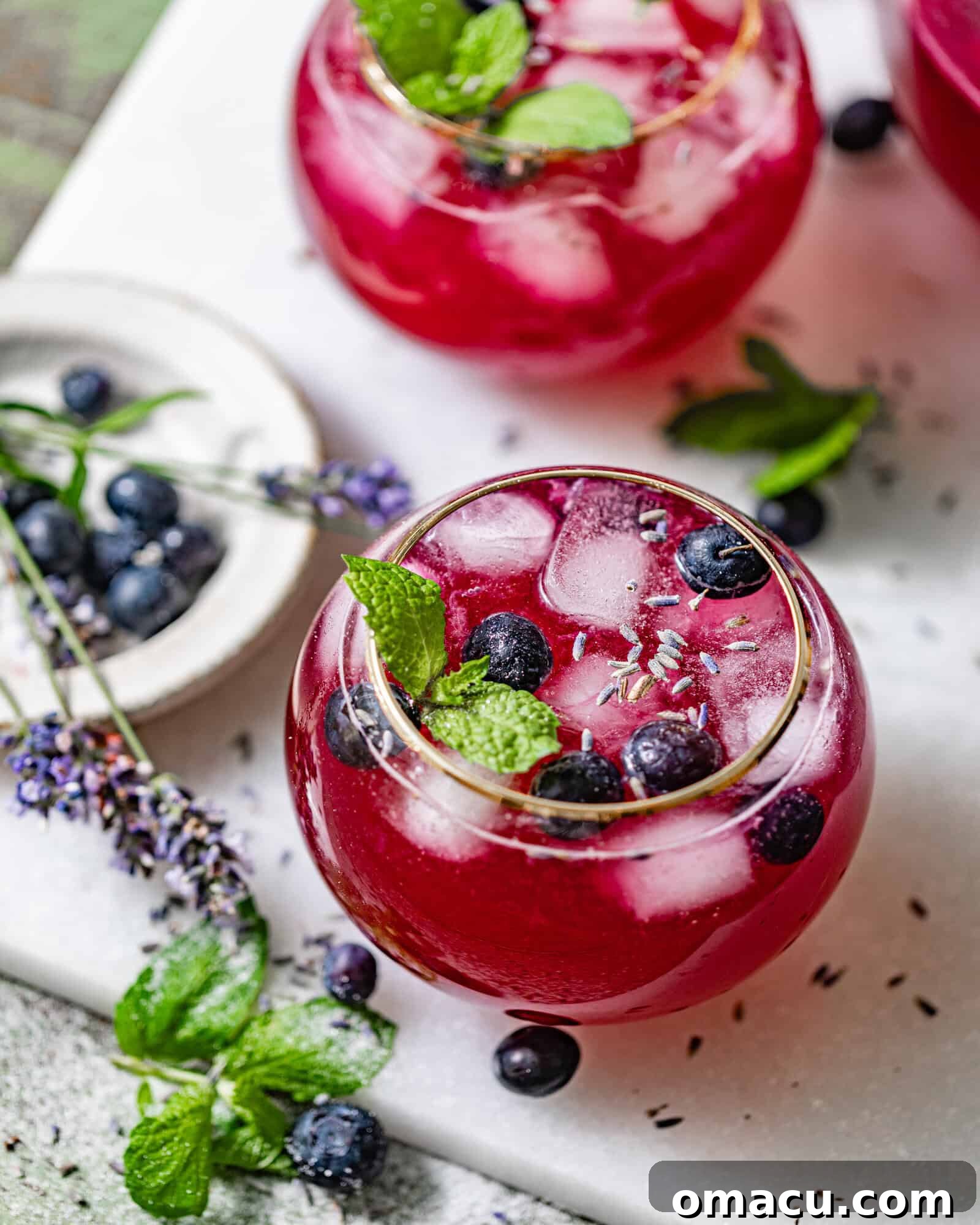 A frosty glass of vibrant Blueberry Lemonade, brimming with ice cubes, fresh blueberries, and delicate mint leaves, sparkling enticingly in the sunlight.