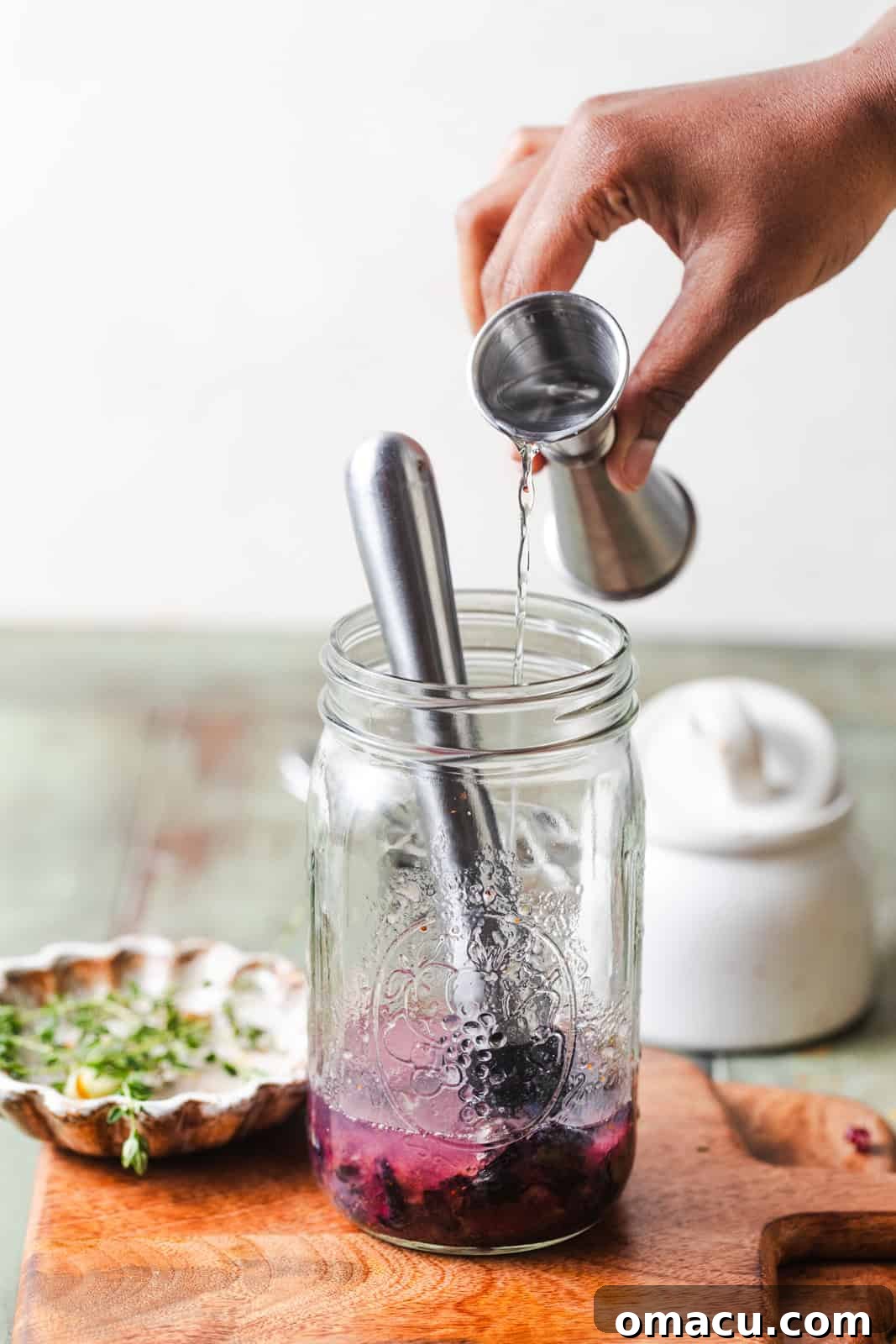 A hand expertly pouring clear vodka into a large mason jar, which already contains a vibrant mixture of muddled blueberries and sweet jam, ready for the next step of cocktail creation.