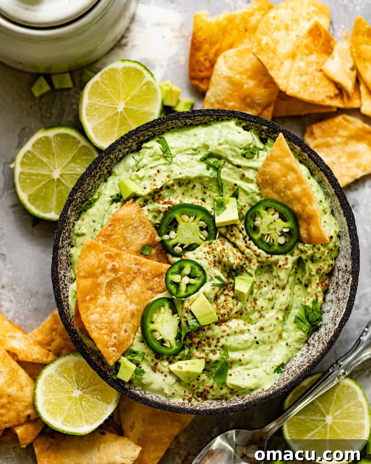 A vibrant bowl of creamy avocado crema, garnished with fresh sliced jalapeños, a sprinkle of Tajín seasoning, and crispy tortilla chips on top, ready to be served.