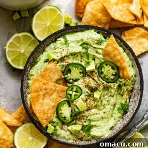 A close-up shot of creamy avocado crema in a bowl, garnished with sliced jalapeños, a dash of Tajín, and surrounded by crispy tortilla chips.