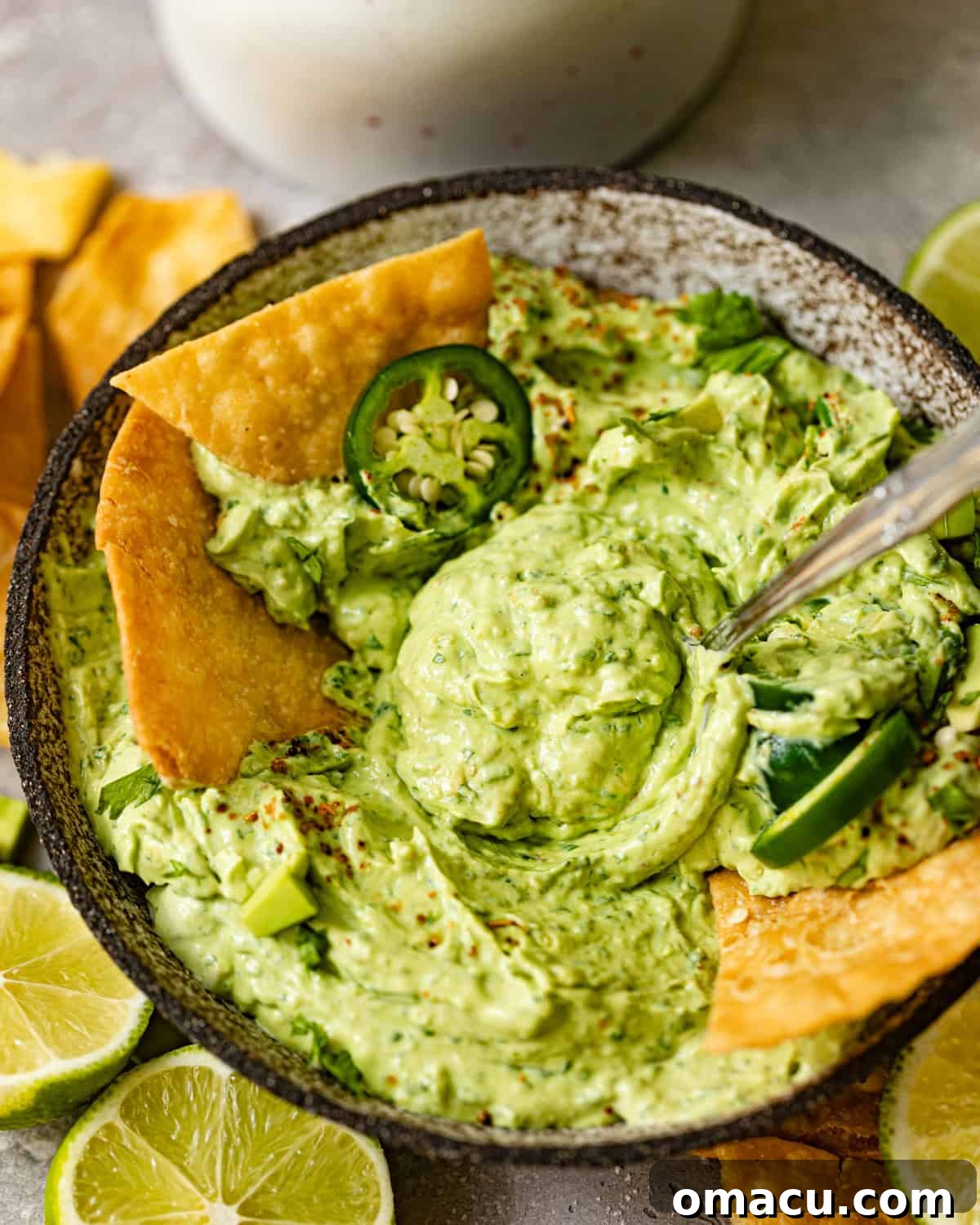 A bowl of creamy avocado crema, adorned with a spoon and garnished with fresh cilantro and sliced jalapeños, ready for serving.