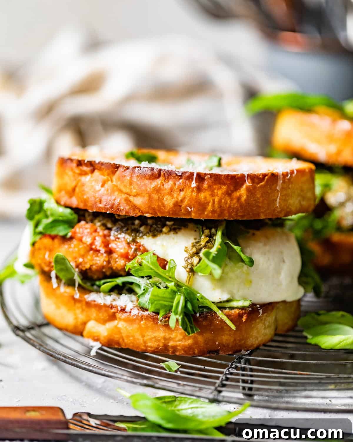 A beautifully plated chicken parmesan sandwich, sliced in half to reveal its layers, resting on a wire rack with fresh basil and grated parmesan nearby.