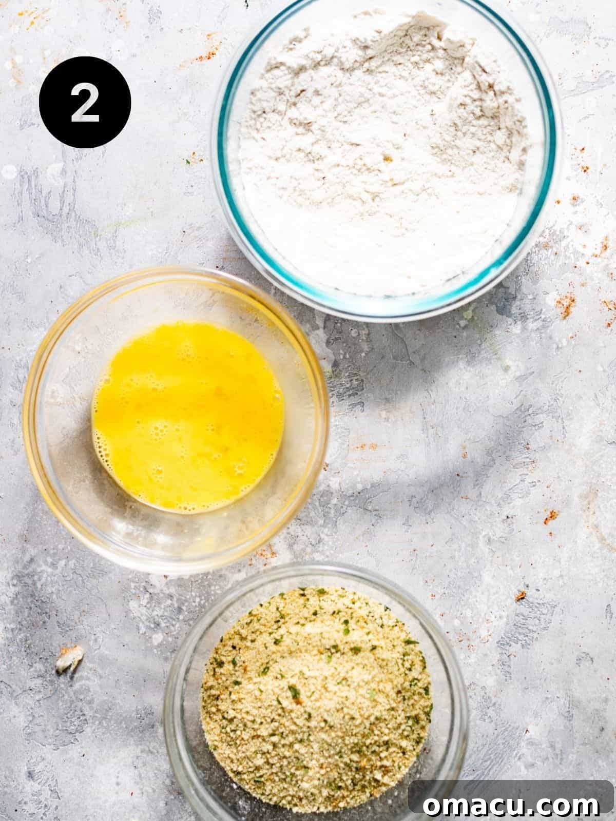 Three shallow bowls arranged as a breading station: one with all-purpose flour, one with beaten egg and water, and one with a mixture of panko breadcrumbs, Parmesan, and seasonings.