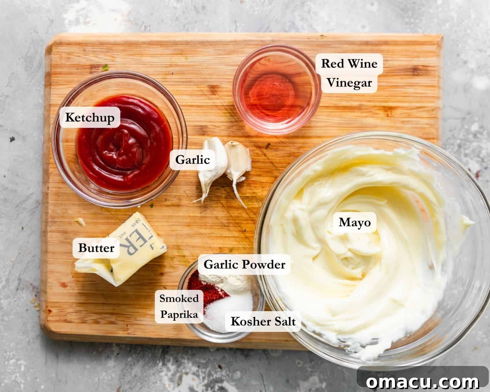 All the ingredients needed to prepare the creamy Yum Yum sauce topping, neatly arranged on a rustic cutting board. Items include a jar of mayonnaise, a bottle of ketchup, red wine vinegar, a stick of butter, fresh garlic cloves, smoked paprika, garlic powder, and a sprinkle of kosher salt.
