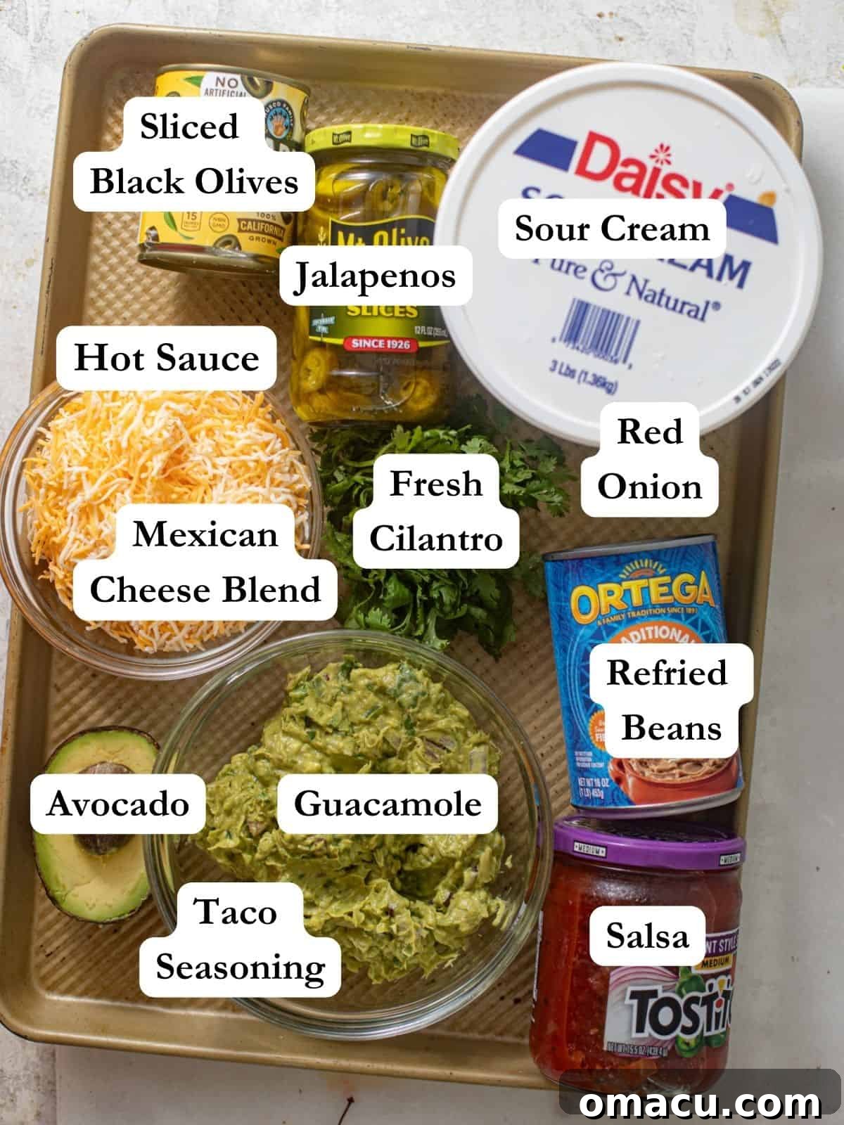 All the fresh ingredients for a five layer dip laid out on a baking sheet, including sour cream, refried beans, Mexican cheese blend, olives, jalapenos, guacamole, salsa, cream cheese, taco seasoning, avocado, and red onion, highlighting the vibrant colors and fresh quality.