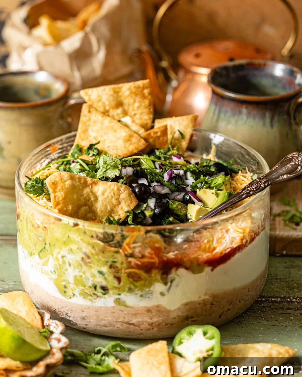 A beautifully arranged five layer dip in a clear glass bowl, generously topped with fresh cilantro, sliced black olives, finely diced red onion, and surrounded by golden tortilla chips, inviting guests to dig in.