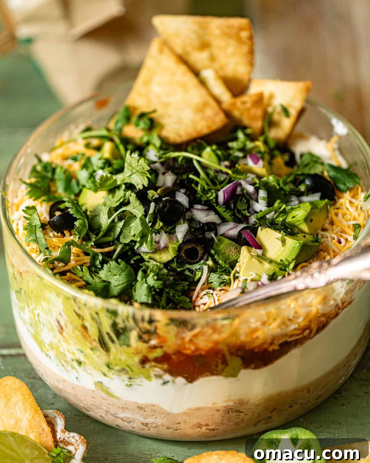 A large glass bowl filled with the colorful layers of five layer dip, with a spoon digging in, showing the texture and freshness of the appetizer.